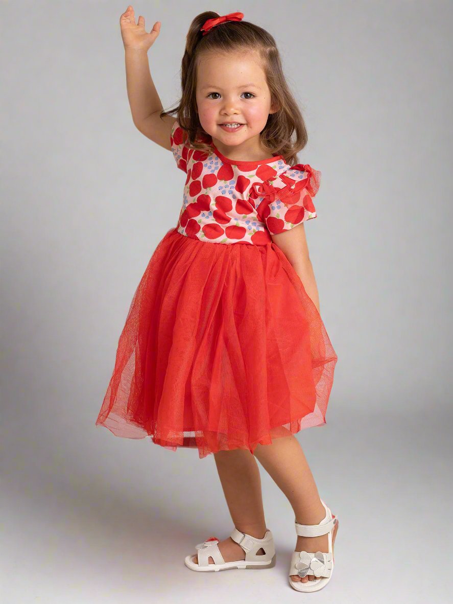 A young girl in the Sweet Apple Ariel Dress, a charming party piece featuring a red and white floral design with a tulle tutu skirt, is smiling and raising her hand. The stretch jersey bodice adds comfort as she stands in her white sandals on a tiled floor against a plain background.