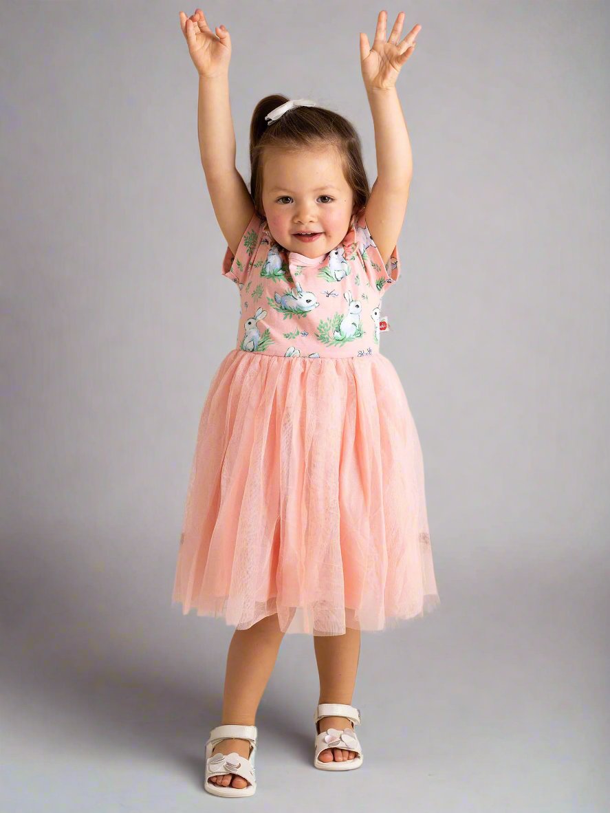 A toddler wearing a Pink Bunnies Ariel Dress from Bunny stands on a tiled floor, raising both arms with a cheerful expression.