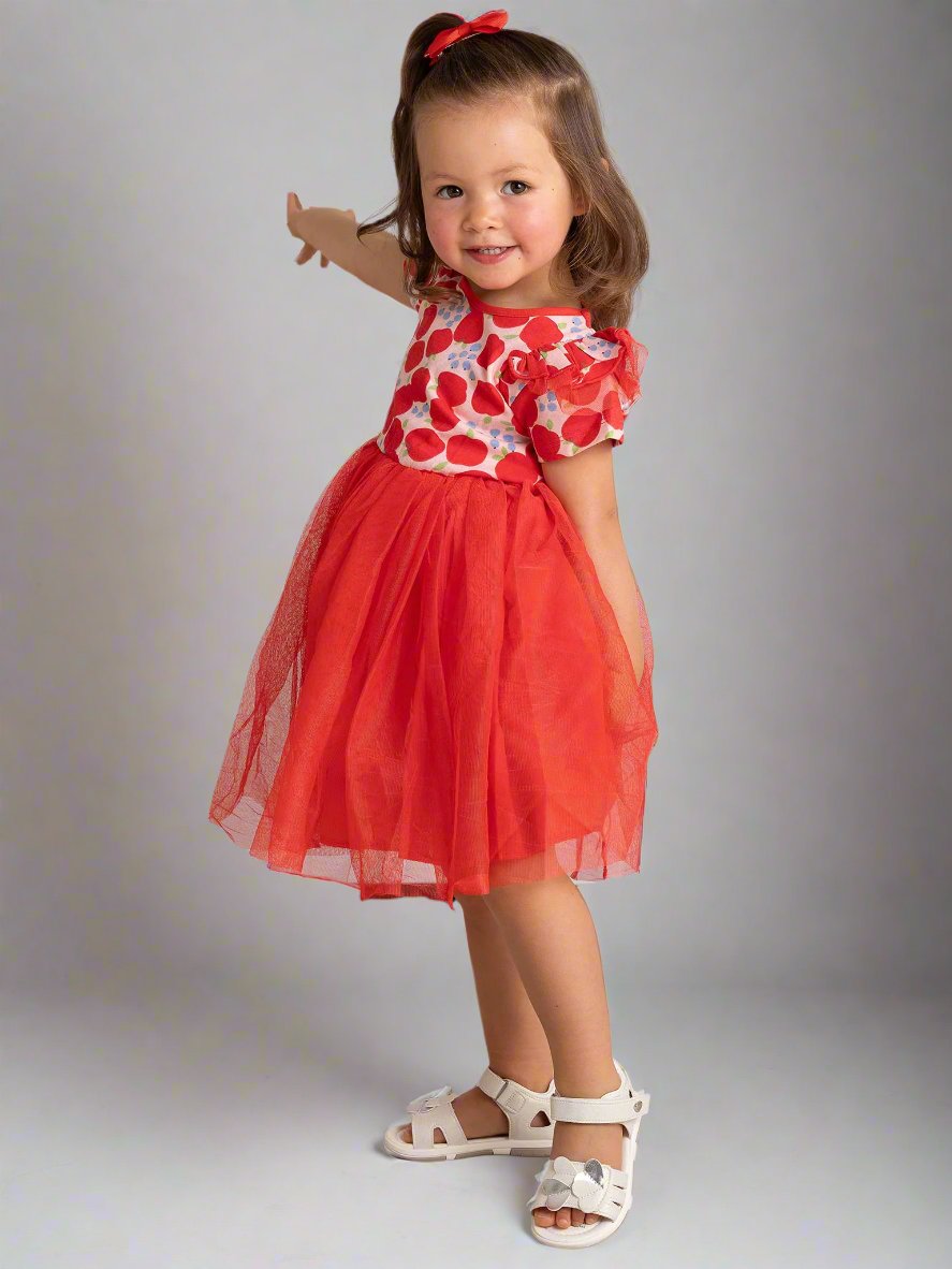 A young girl in the Sweet Apple Ariel Dress, featuring a delightful tutu skirt, stands on a tiled floor, smiling and posing with one arm extended. Her outfit, perfect as a party piece, adds a playful touch to the charming ensemble.