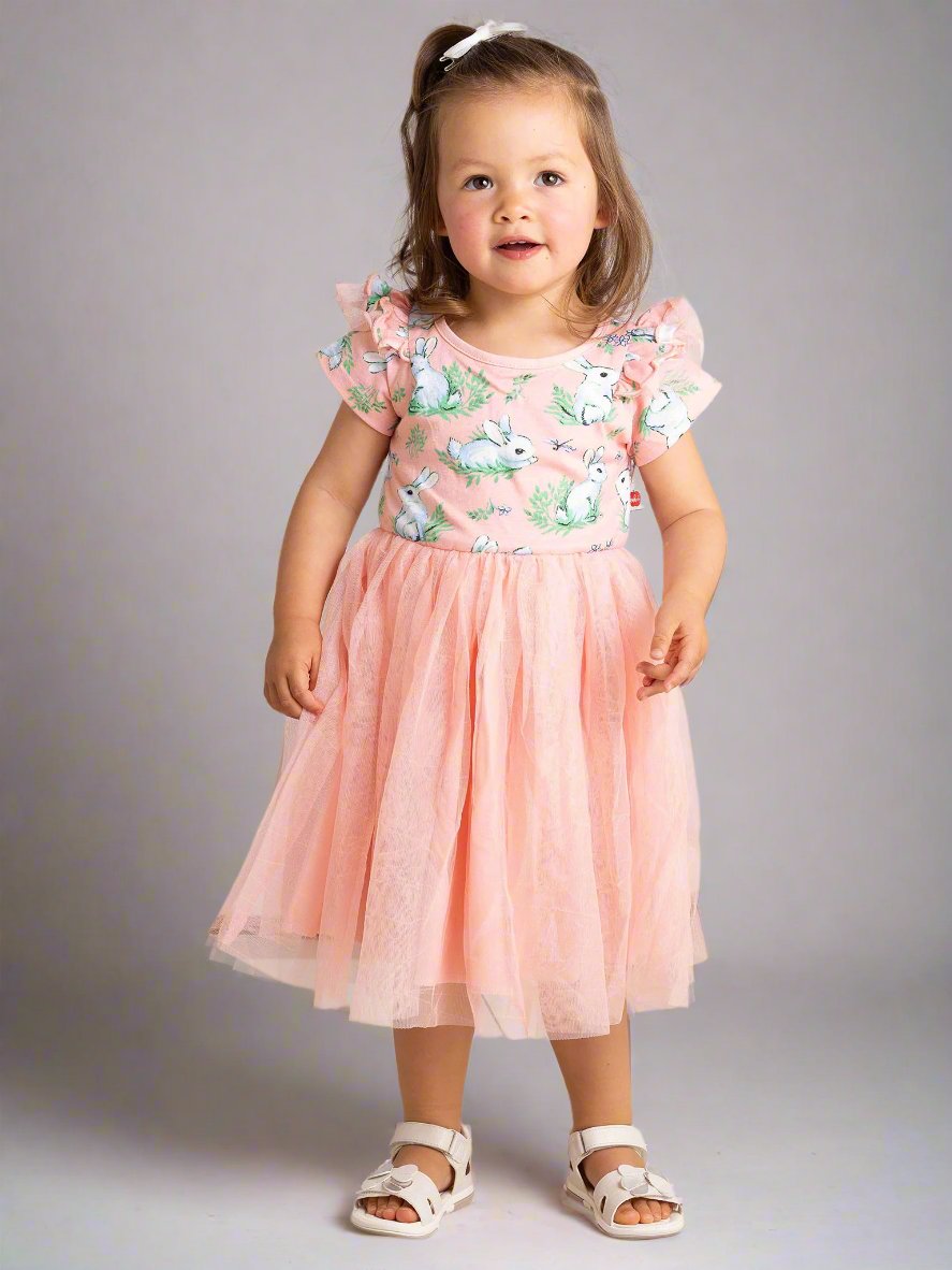 A toddler stands on a polished floor wearing a Bunny Pink Bunnies Ariel Dress in pastel pink adorned with floral patterns, crafted from soft cotton jersey, and paired with white sandals.