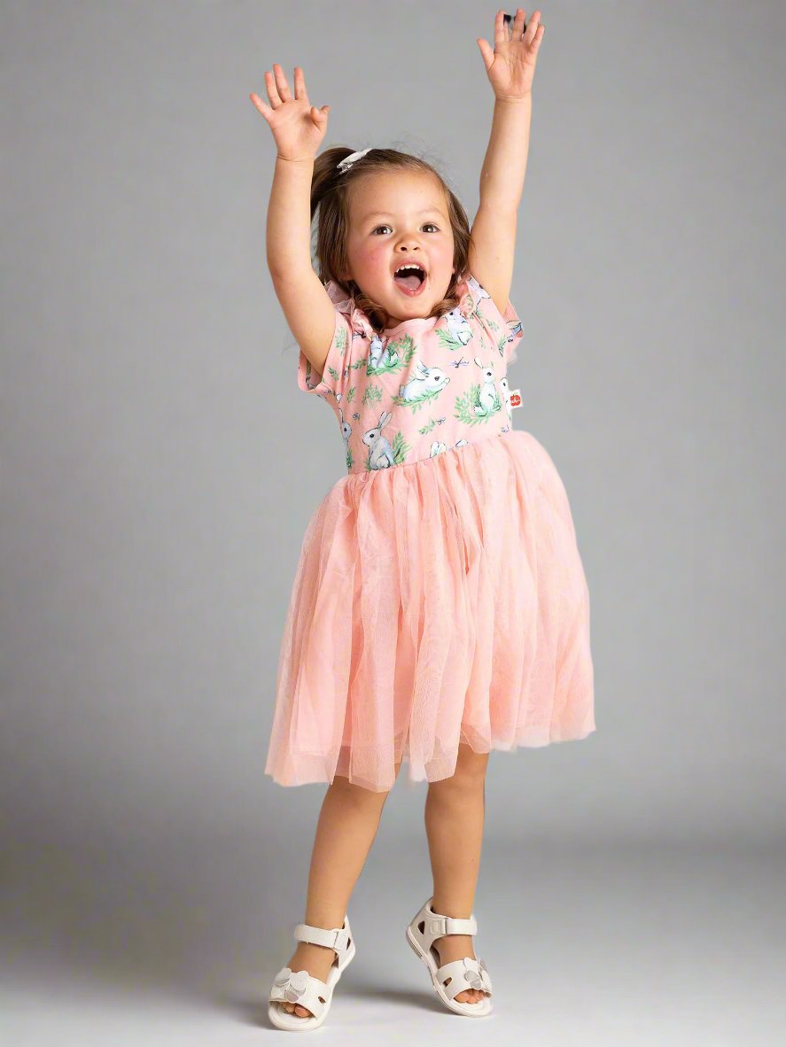 A young child wearing a Pink Bunnies Ariel Dress by Bunny, made of soft cotton jersey, jumps with arms up in front of a backdrop.