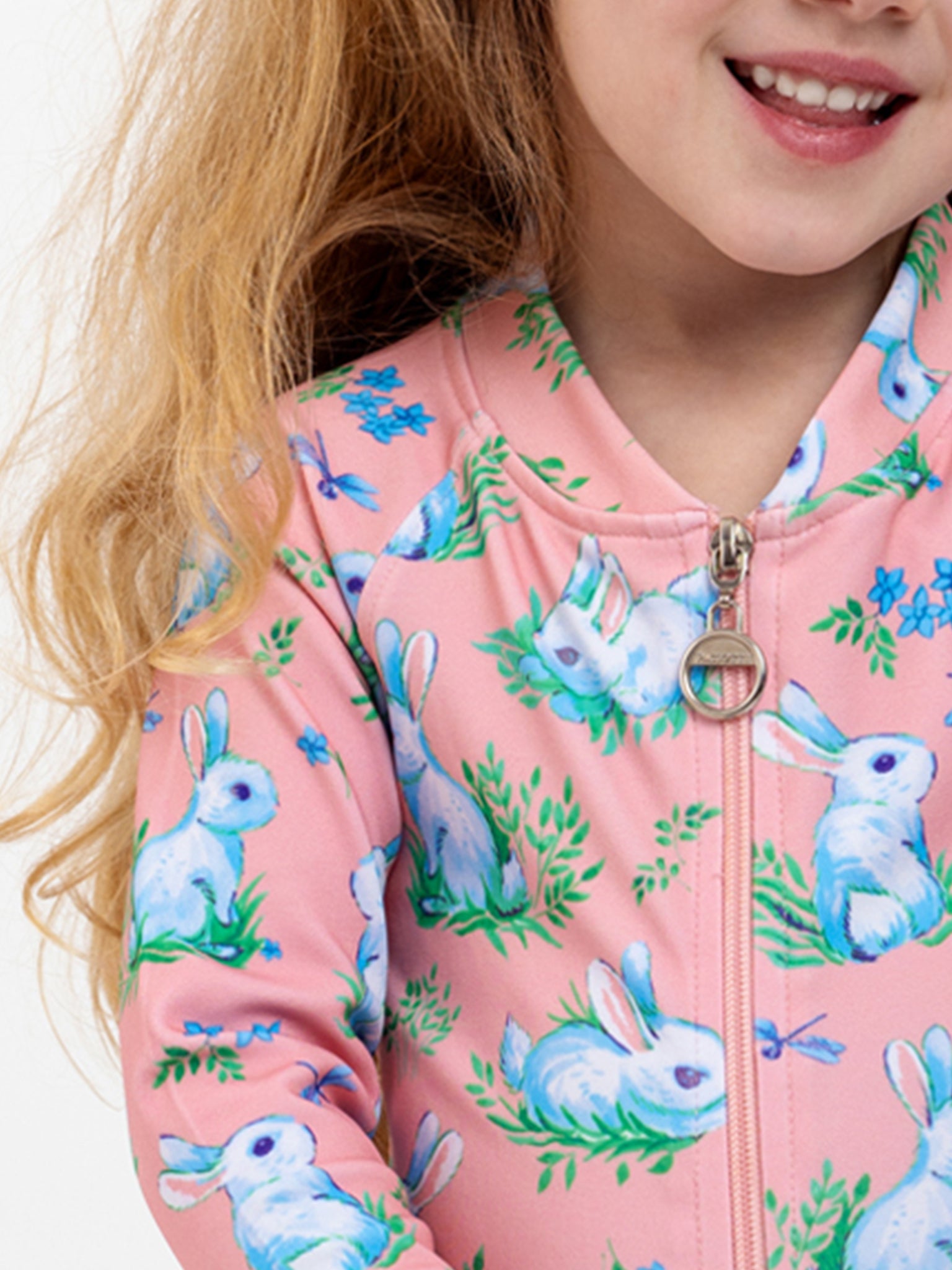 A child in a Pink Bunnies SPF50 Zippy Swimsuit by Bunny, adorned with blue and white rabbit patterns, is smiling gleefully. The swimsuit, reminiscent of a pink jacket, features a zippered front with a circular zipper pull, adding charm to the little mermaid's vibrant day.