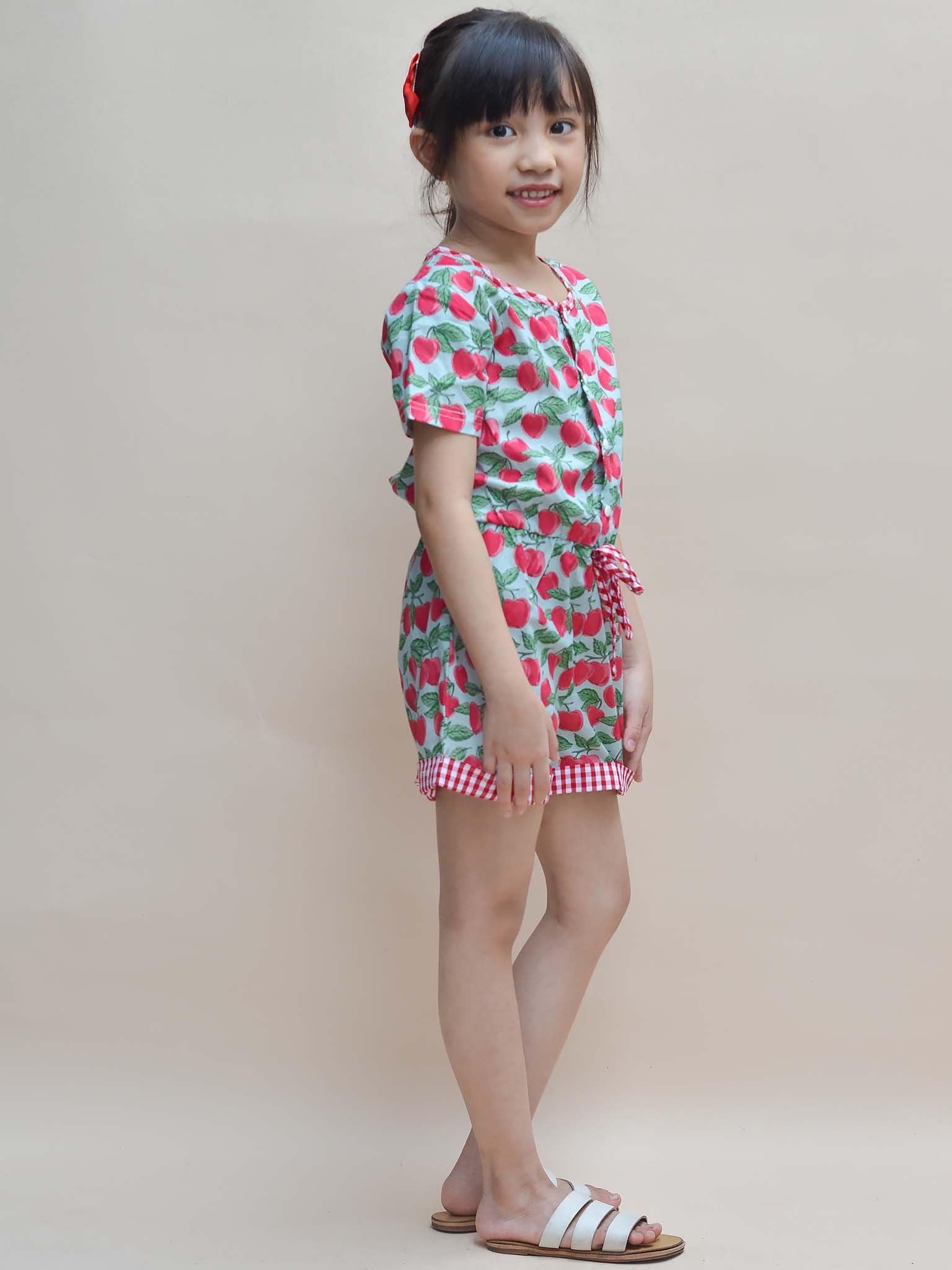 A young girl named Maisy stands smiling in a Blue Cherry Maisy Playsuit, her red bow and sandals adding charm against a neutral beige background, epitomizing delightful girls' clothes.