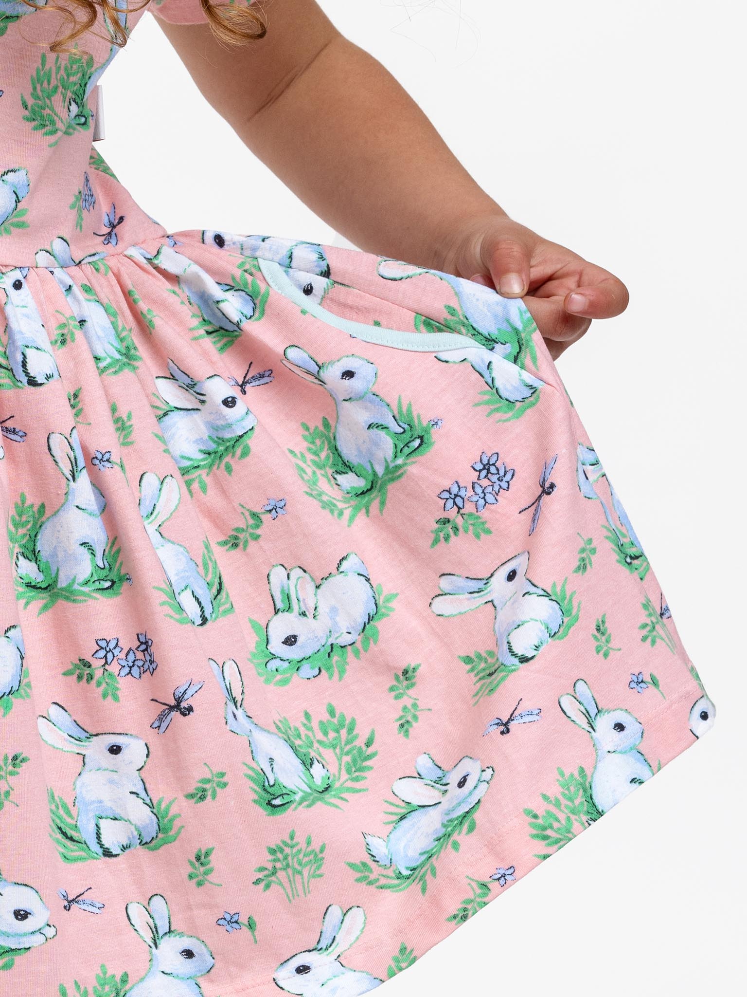 A child is holding the hem of a Pink Bunnies Amelie Dress from Bunny, featuring its pink cotton jersey fabric decorated with a charming pattern of white rabbits and blue flowers.