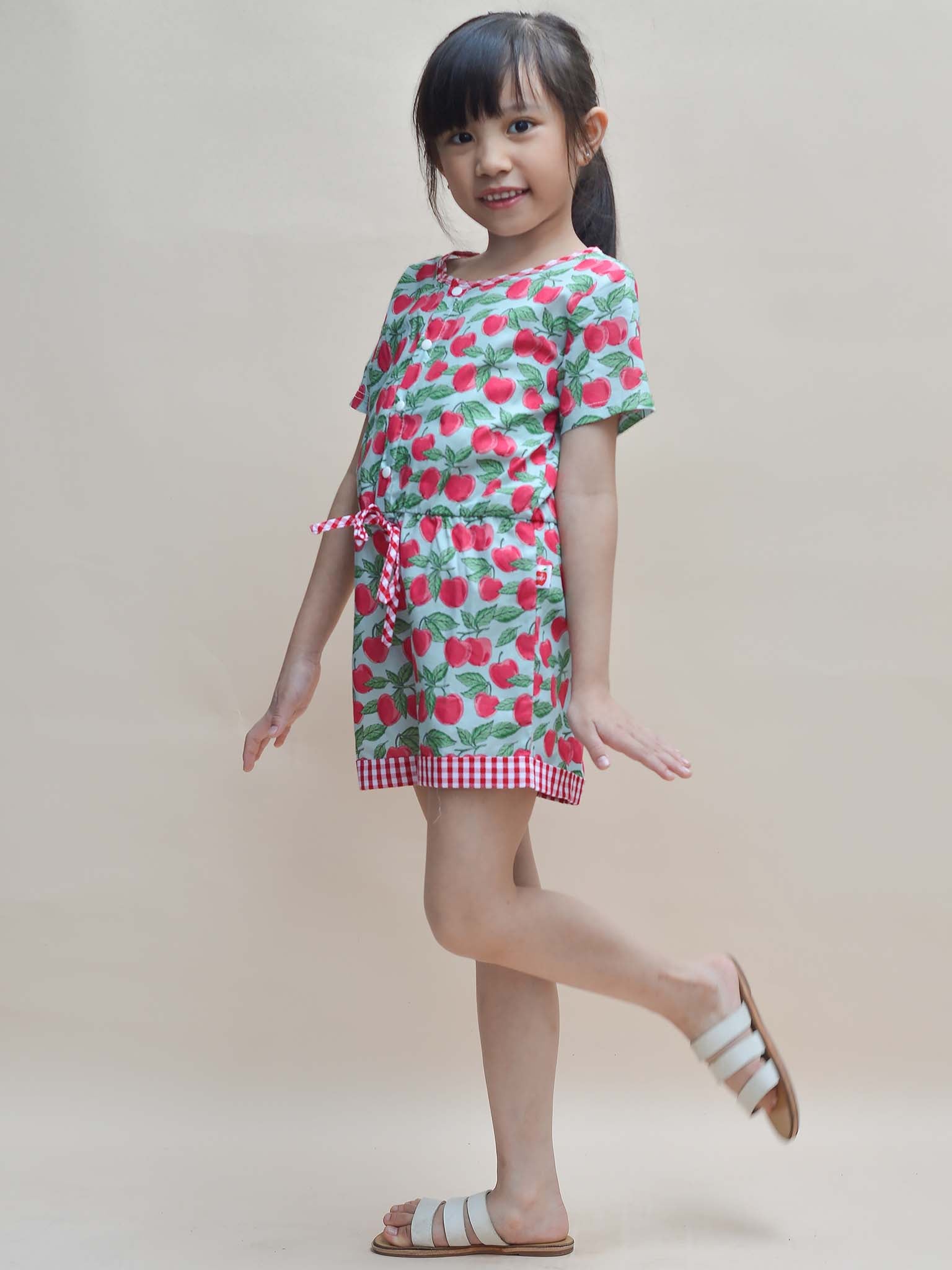 Maisy beams as she gracefully poses in her Blue Cherry Maisy Playsuit and white sandals, showcasing the charming essence of girls' clothes against a neutral background.