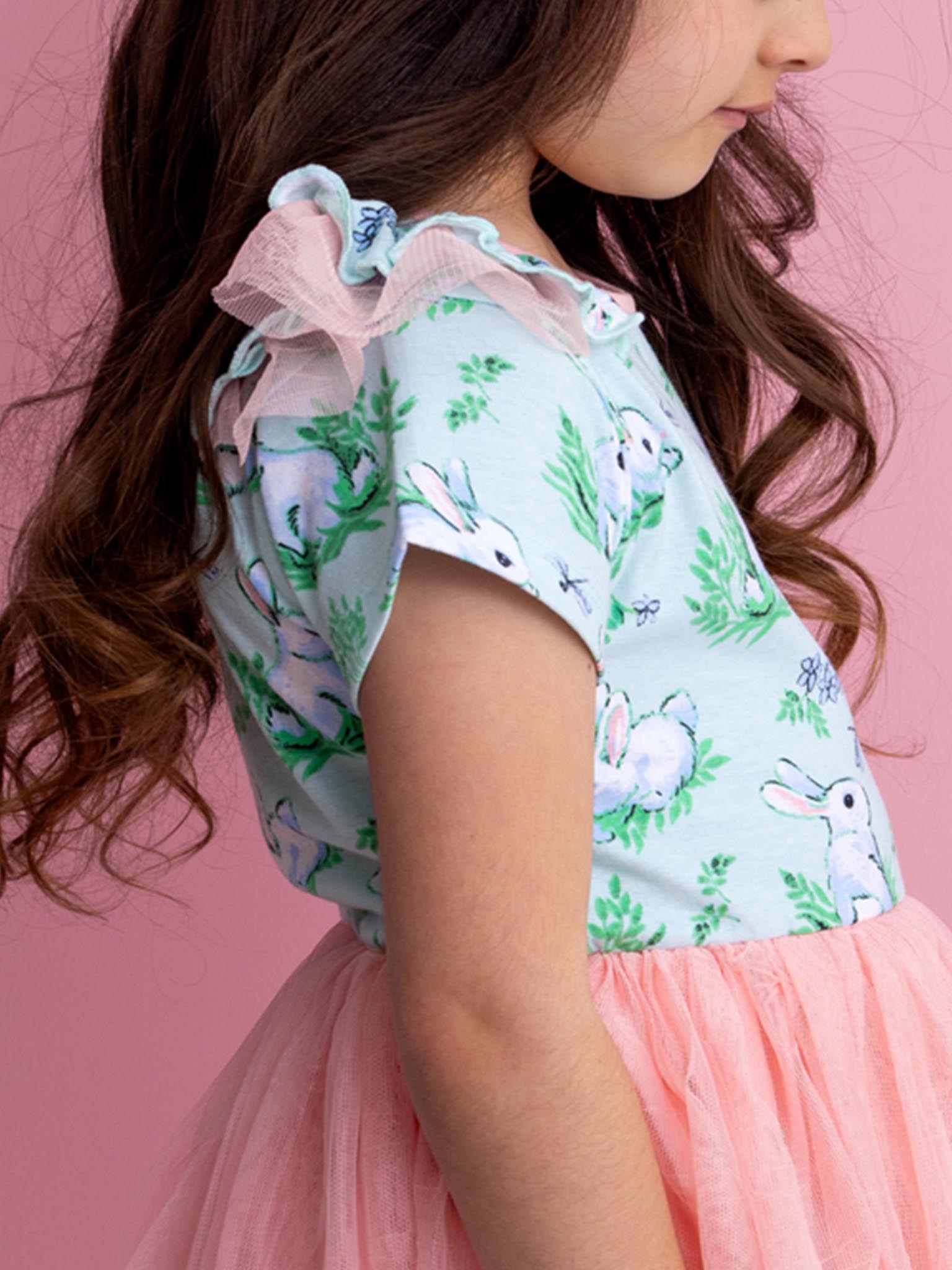 A young girl wearing the Bunny brand's Blue Bunnies Ariel Dress, featuring charming bunny designs and a pink tulle skirt, stands against a pink background.