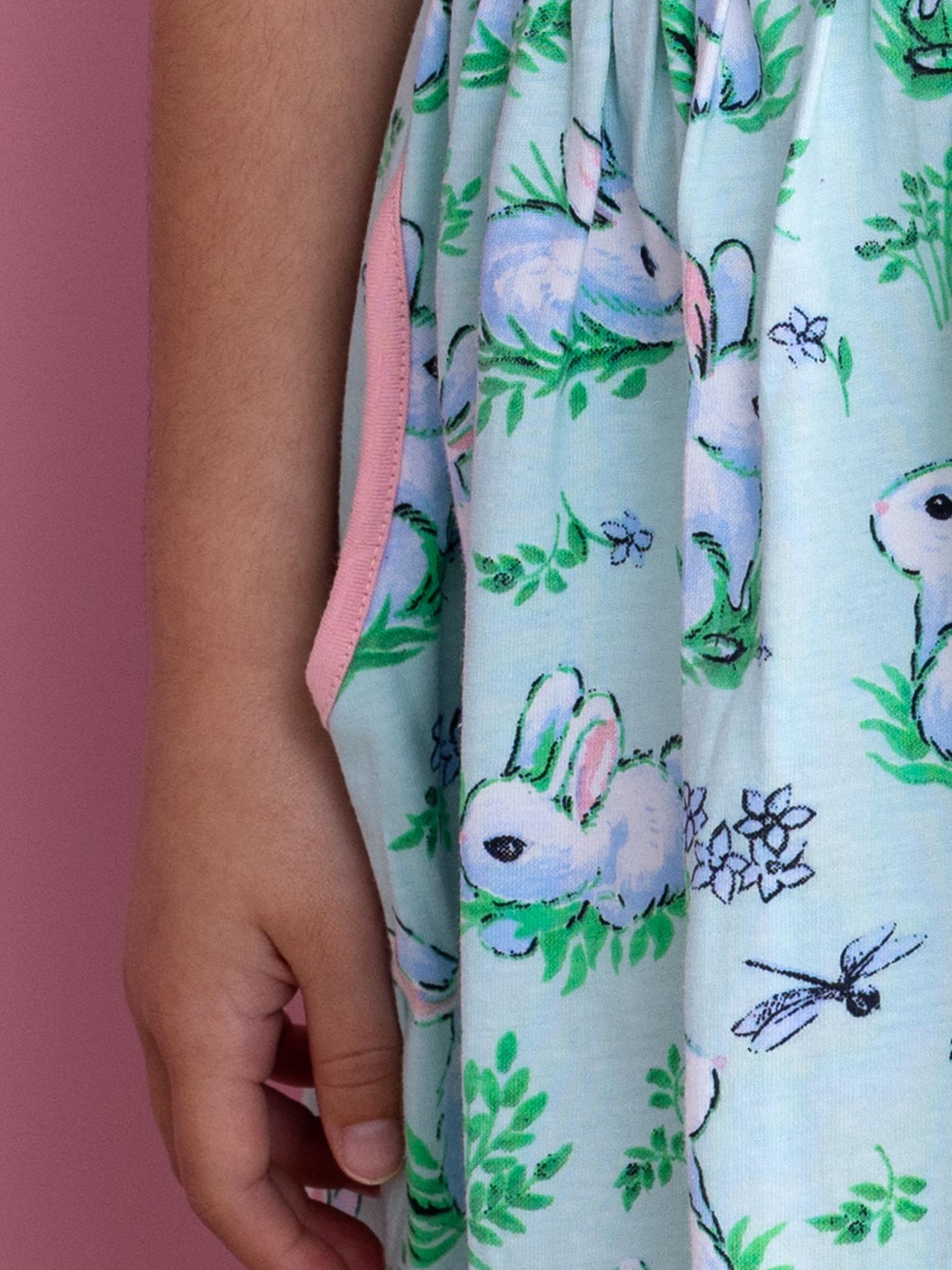 A child's hand gently rests on the Blue Bunnies Amelie Dress by Bunny, featuring a charming rabbit and floral pattern set against a soft pink background. Made from 100% cotton jersey, its hand-printed design offers both comfort and whimsy.