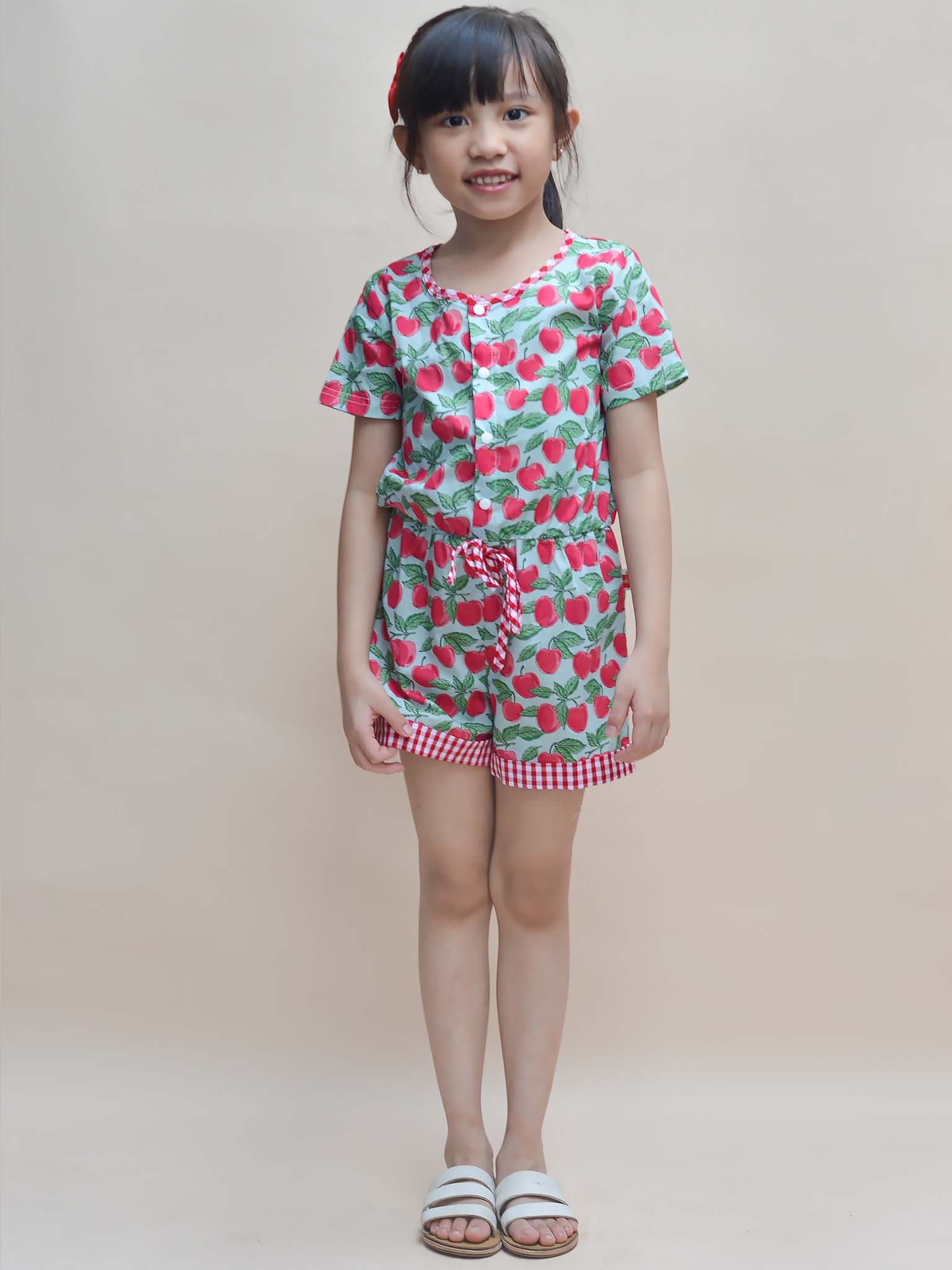 A young girl stands against a beige background, wearing a Blue Cherry Maisy Playsuit and white sandals, embodying the whimsical charm of Blue Cherry's delightful girls' clothes.