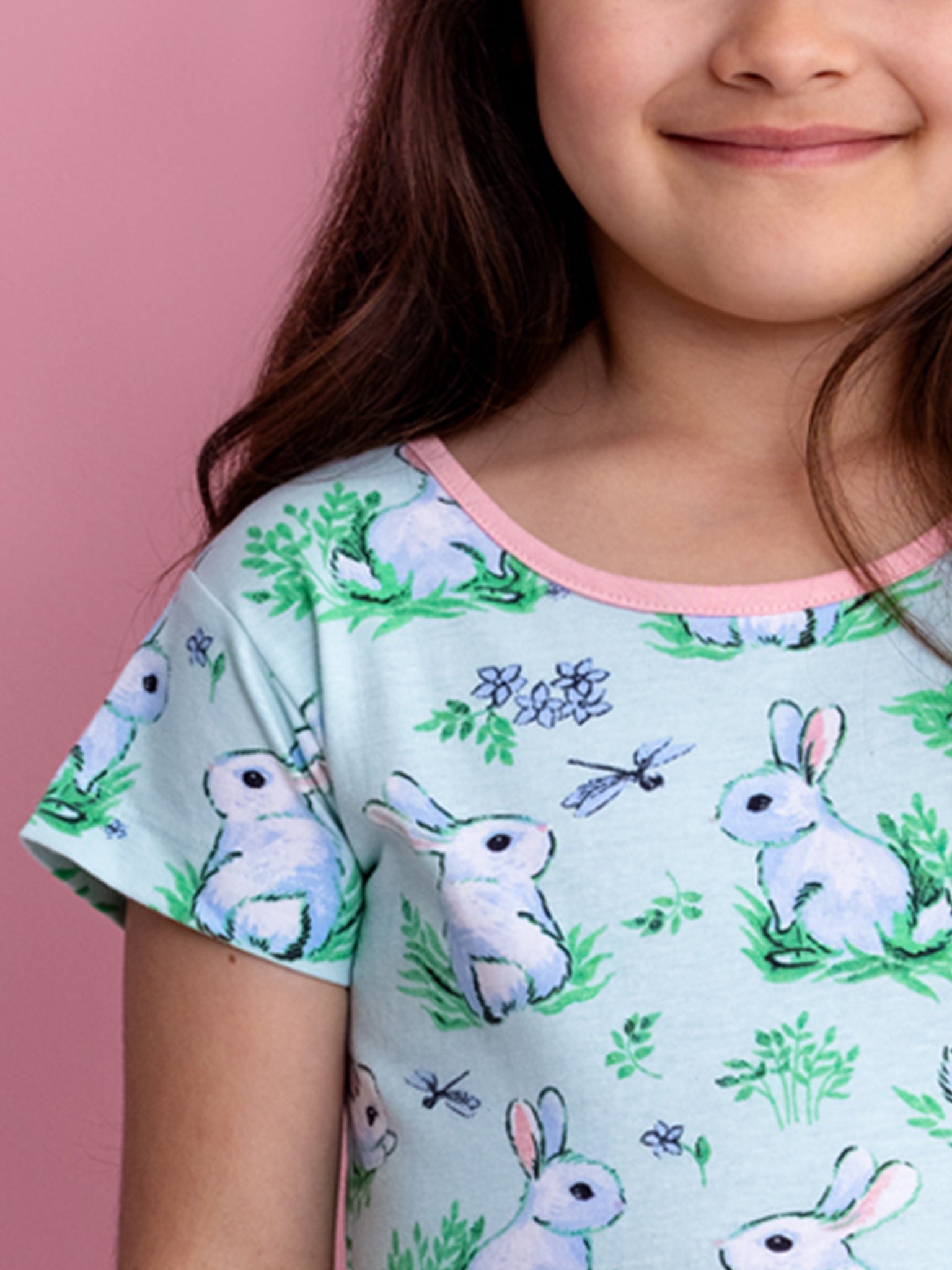 A child in the Blue Bunnies Amelie Dress from Bunny, crafted from 100% cotton jersey with a bunny pattern, sits against a pink background.