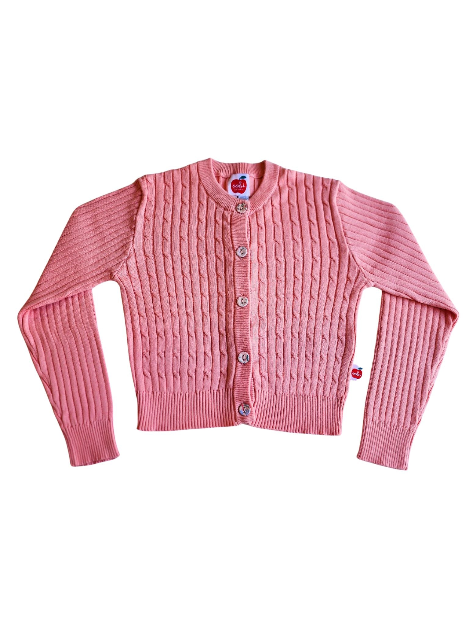 Introducing the Maxine Pink Icing Cardigan by Knitwear: a pink knitted masterpiece made from 100% cotton cable knit, featuring a charming button-up front with fabric-covered buttons and a small red apple emblem on the bottom right hem.