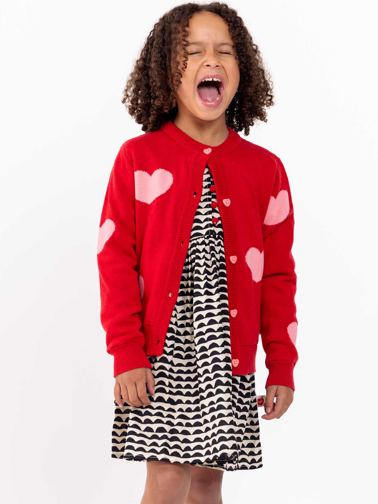 A young girl in a Knitwear Maxine Red with Pink Hearts Cardigan and a black and white wavy dress, standing and yawning with her eyes closed.