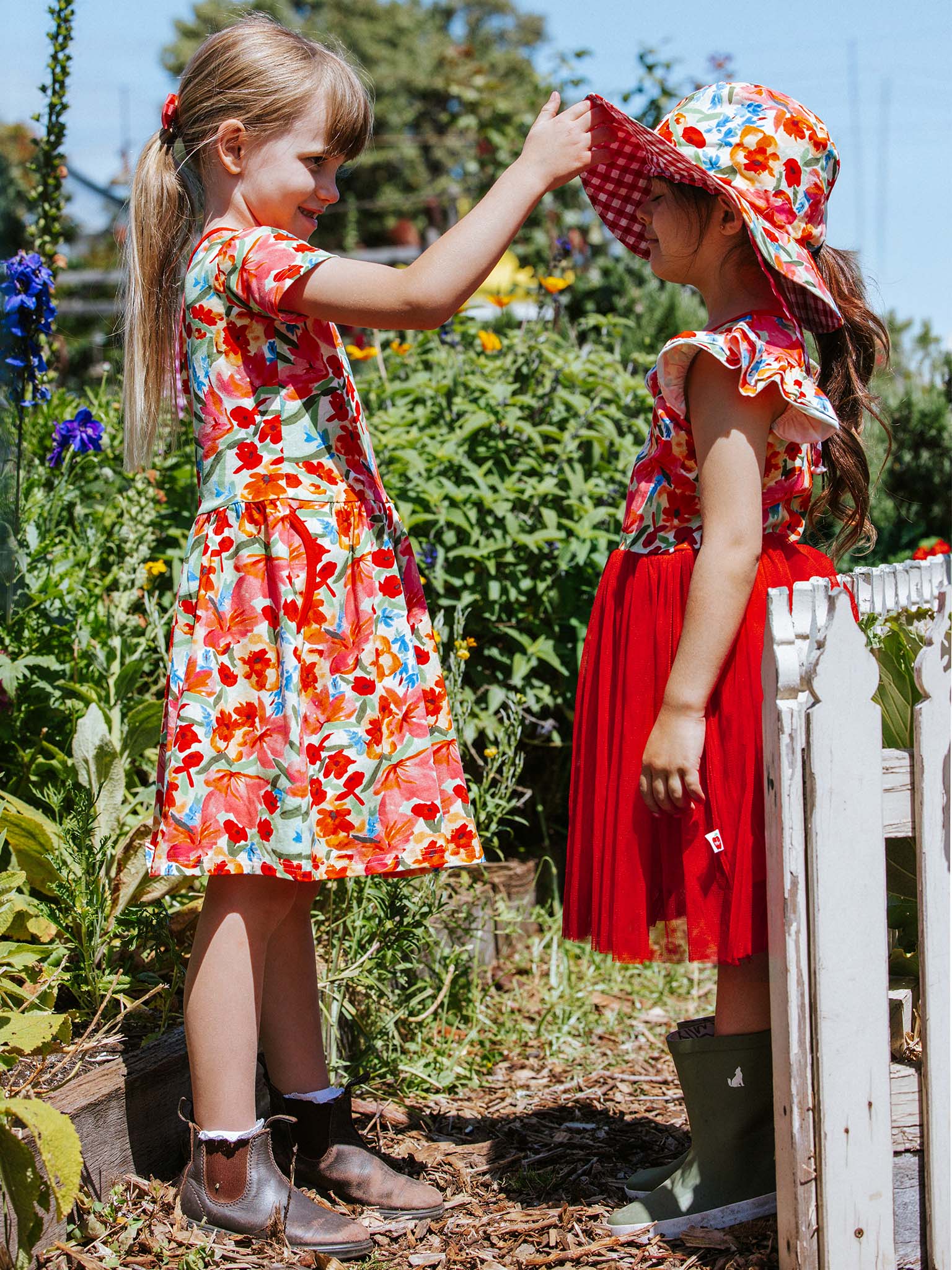 Two young girls in vibrant floral outfits from Wild Roses, dressed in the whimsical Ariel Dress, stand in a garden. One adjusts the other's hat, adding a charming touch to their enchanting playtime scene.