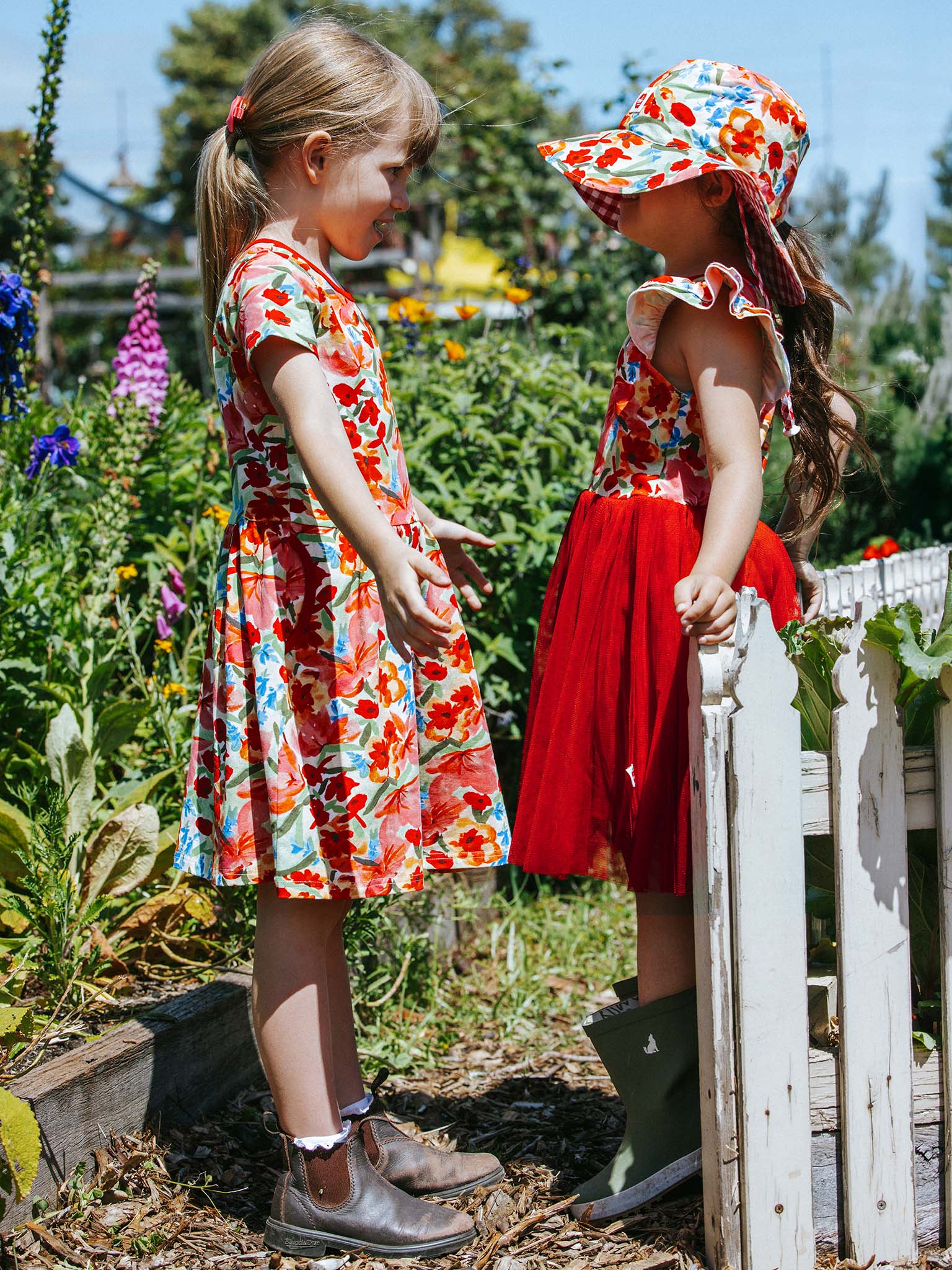 In a garden lush with greenery and wild roses, two young girls in Wild Roses Amelie Dresses, crafted from stretchy jersey fabric adorned with floral patterns, stand facing each other.