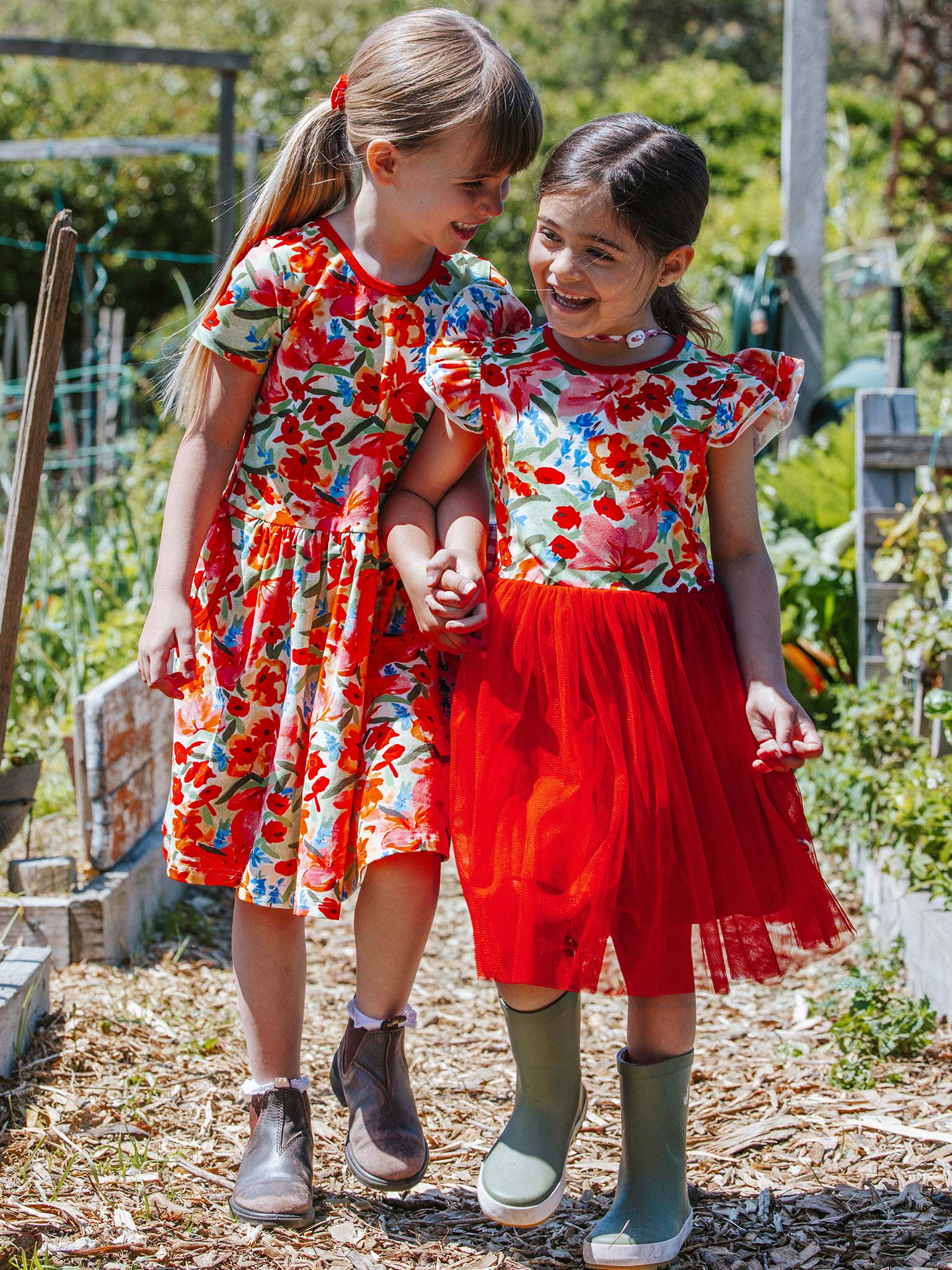 Two young girls stroll along a garden path, wearing vibrant Wild Roses Ariel Dresses and boots, smiling at each other.