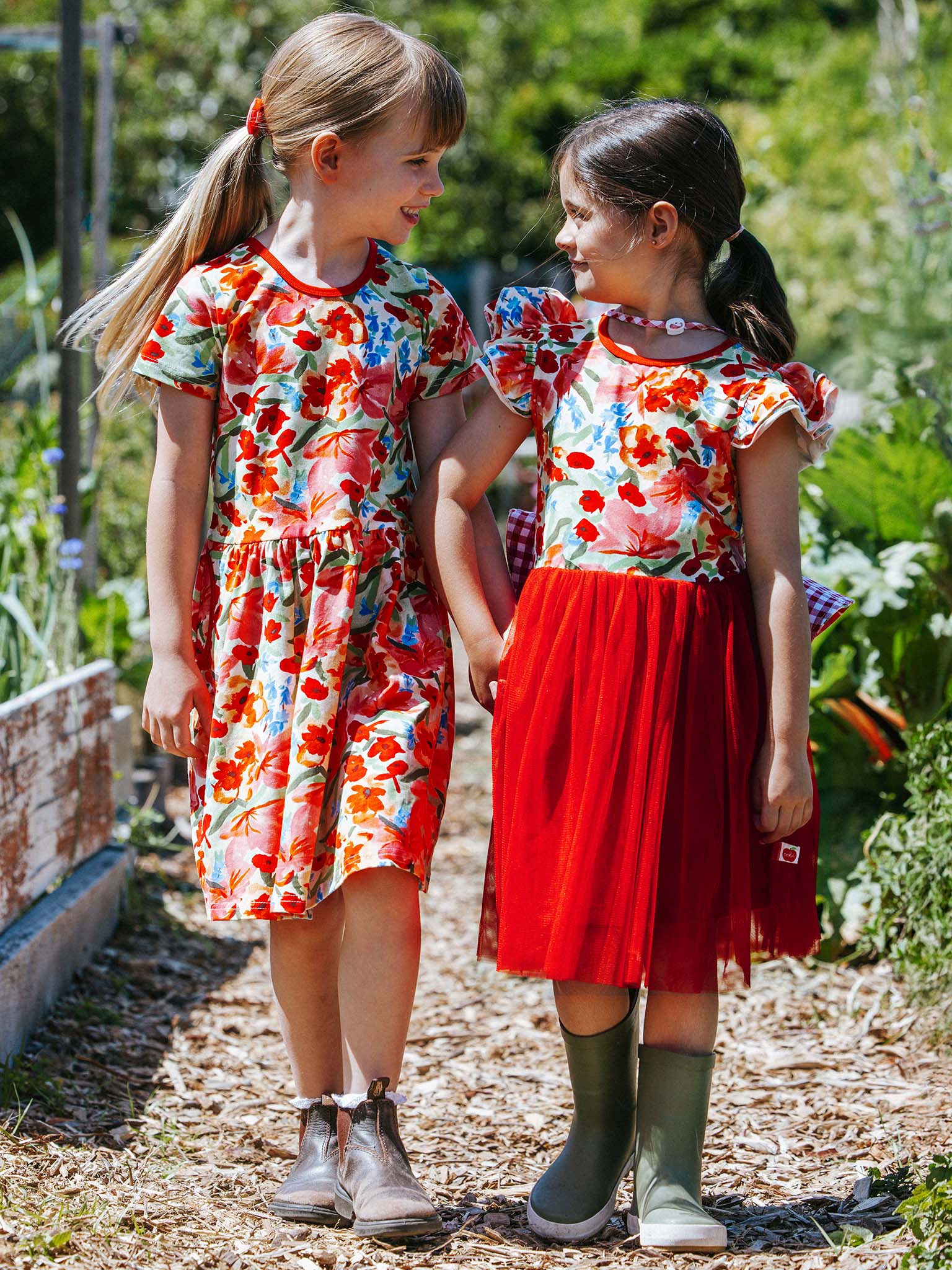 Two young girls in beautiful Wild Roses Ariel Dresses, showcasing the delightful Wild Roses print, stand on a garden path. Holding hands and smiling at each other, their ponytails sway gently in the breeze.