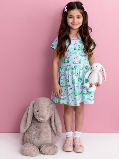 A young girl in a Blue Bunnies Amelie Dress by Bunny, crafted from 100% cotton jersey, holds a stuffed rabbit and stands beside a large plush rabbit against a pink background.