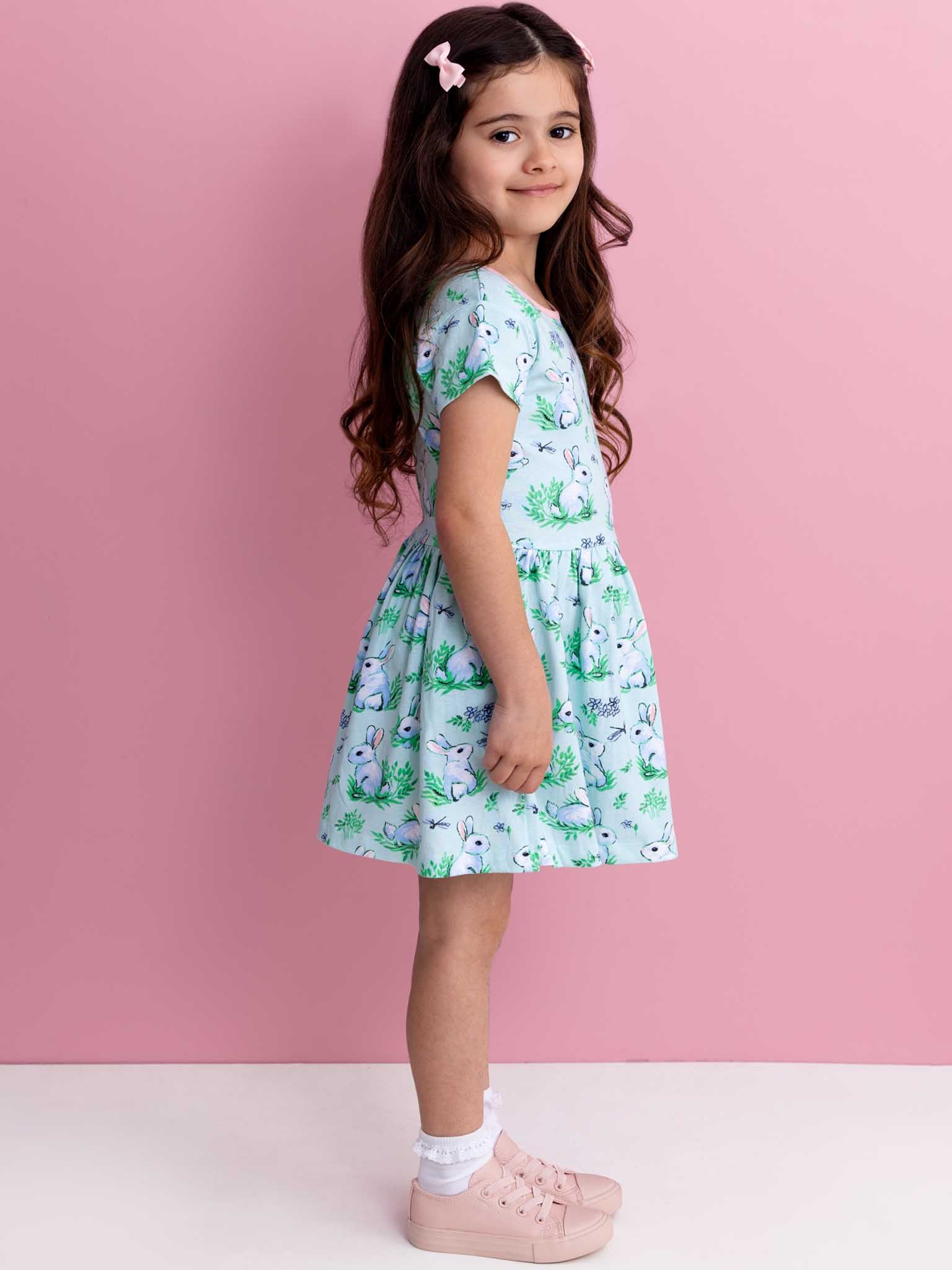 A young girl with long dark hair is dressed in a Blue Bunnies Amelie Dress by Bunny, paired with pink shoes, as she stands sideways against a pink background.