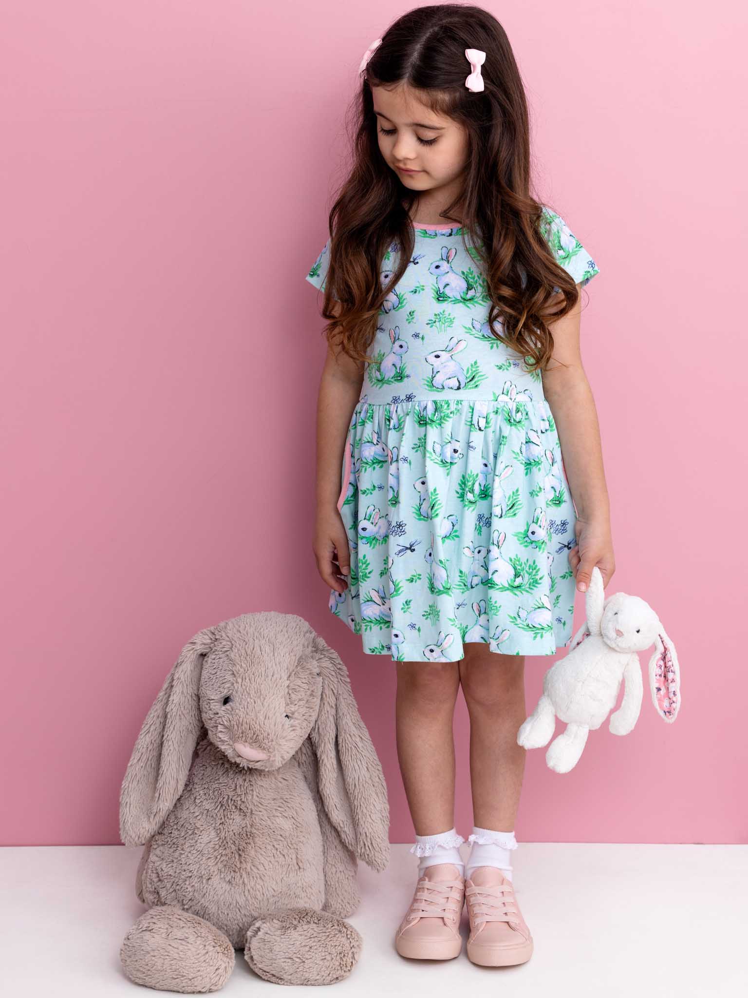 A young girl in a Blue Bunnies Amelie Dress by Bunny, made of 100% cotton jersey, stands on a pink backdrop holding a white stuffed bunny, with a larger gray plush companion resting on the floor beside her.