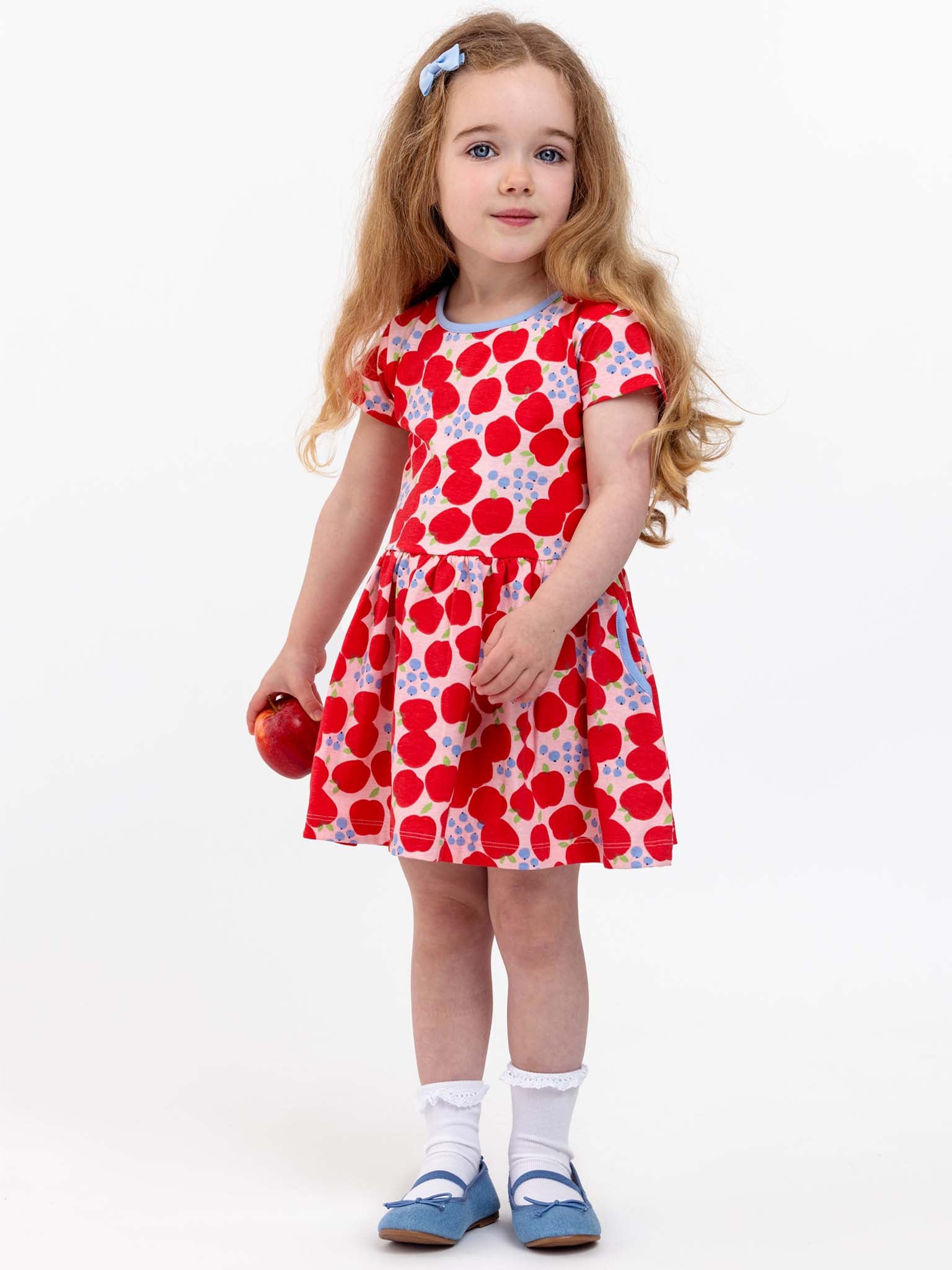 A young girl with long hair stands holding a red apple. She is dressed in the Sweet Apple Amelie Dress, featuring white and blue patterns inspired by the Sweet Apple brand. She completes her look with white socks, blue shoes, and a blue bow in her hair.