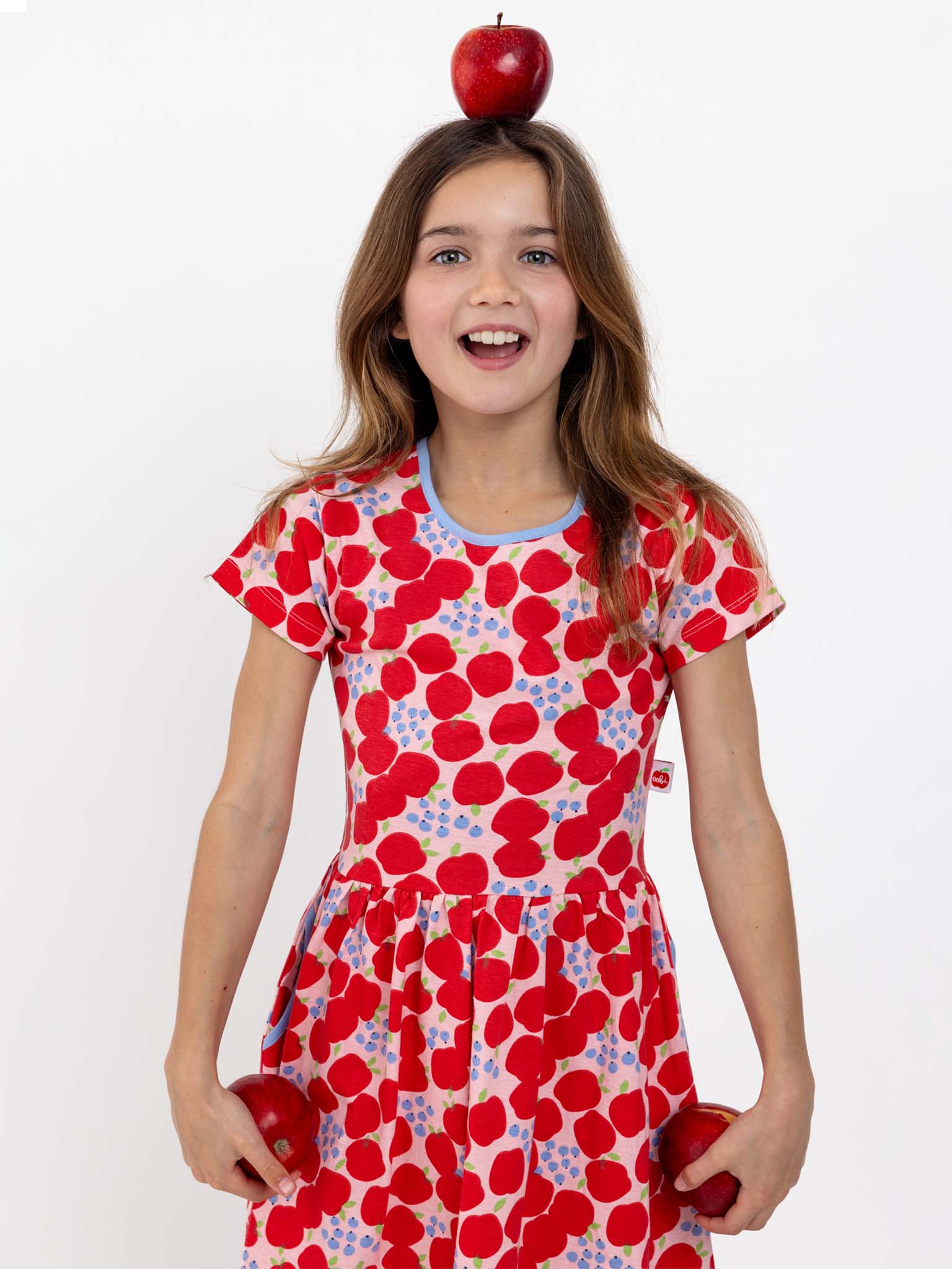 A girl with long hair wears the Sweet Apple Amelie Dress, adorned with a charming red and pink heart pattern. She balances a red apple on her head and holds a red apple in each hand, standing against a plain white background.