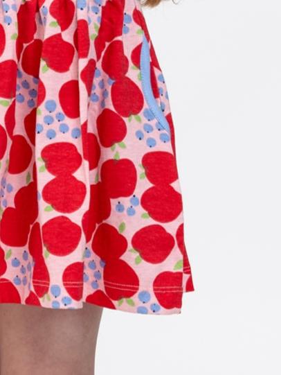 Close-up of a child wearing the Sweet Apple Amelie Dress from Sweet Apple, adorned with a red Sweet Apple print and blue dots. The pink dress, made from stretchy jersey fabric, features a blue pocket on the side. Perfect for girls' dresses collections.