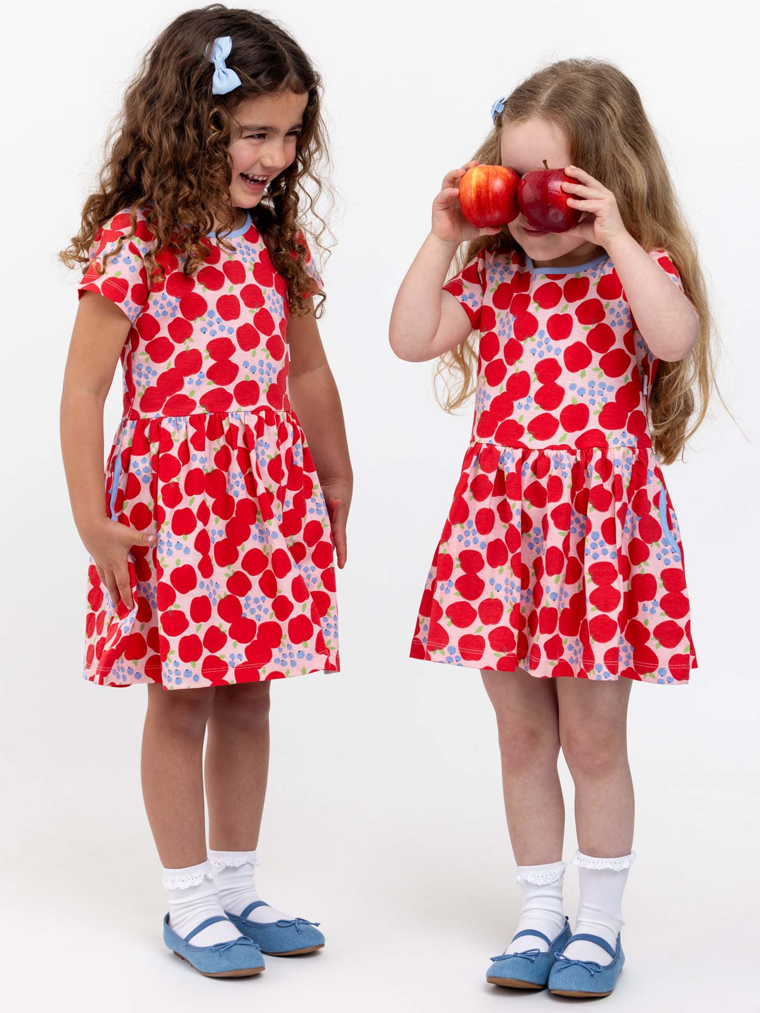 Two young girls in matching Sweet Apple Amelie dresses, which feature a delightful red and white print, paired with blue shoes. The girl on the left is smiling, while the girl on the right playfully holds two apples over her eyes. Both are standing against a plain background. Baby’s got pockets for added charm!