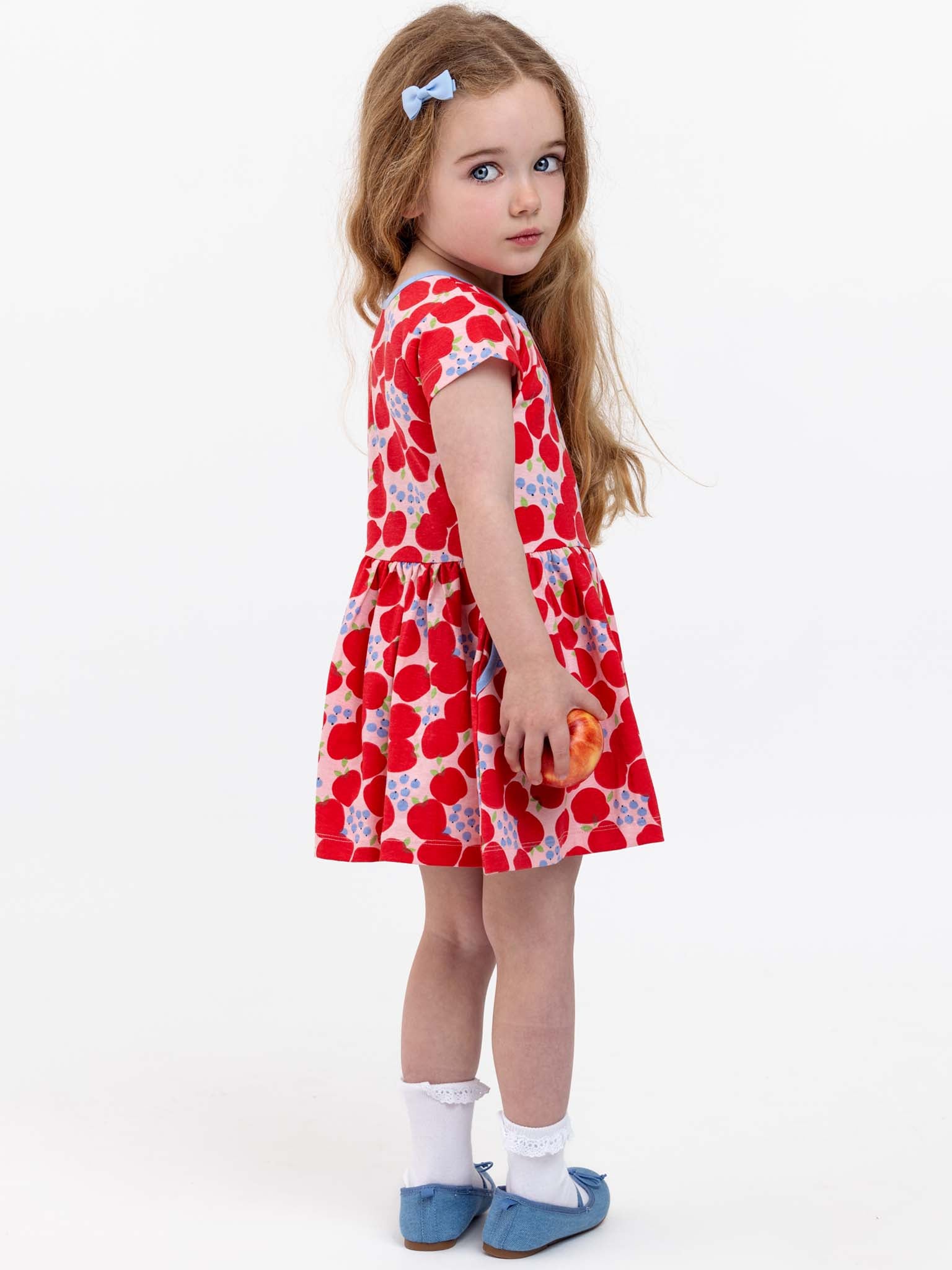 A young girl with long hair and a blue bow stands facing backward, wearing the Sweet Apple Amelie Dress, which features a red and white heart pattern. She also has on white socks and blue shoes. The dress, made of stretchy jersey fabric by the Sweet Apple brand, looks charming as she holds a small toy in her hand.