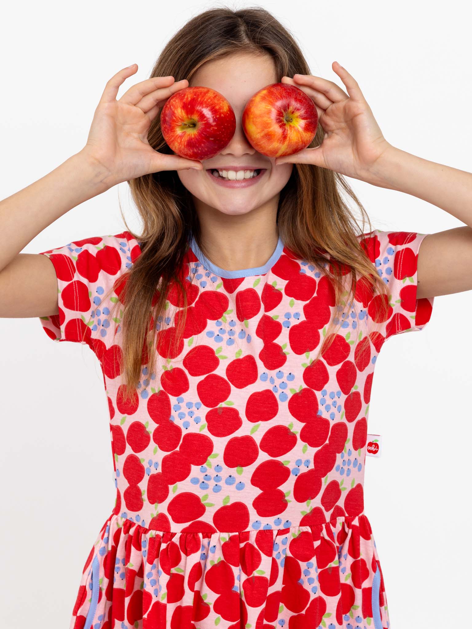 A girl in the Sweet Apple Amelie Dress from Sweet Apple, adorned with a charming red and white polka-dot and sweet apple print, holds two apples over her eyes, smiling.