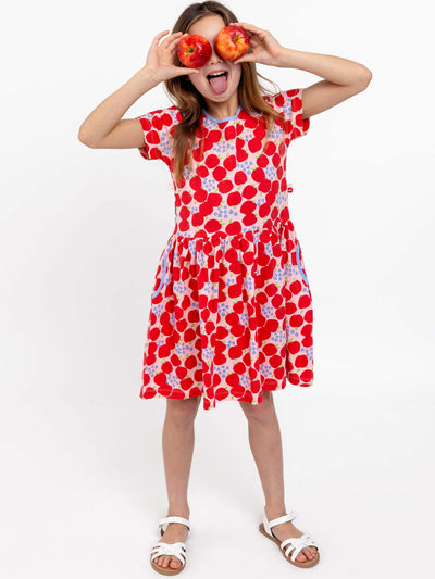 A child in a red, heart-patterned Sweet Apple Amelie Dress, holding two apples in front of their eyes and sticking their tongue out. This playful scene is highlighted by the stretchy jersey fabric of one of the season's cutest girls' dresses from Sweet Apple. White background.