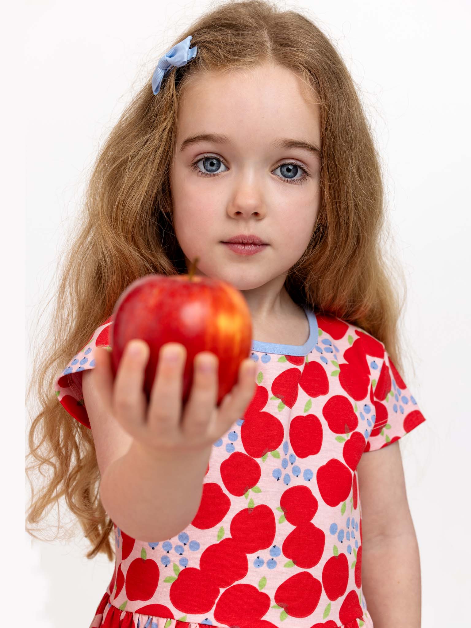 A young girl with long hair and a blue bow holds out a red apple, wearing the Sweet Apple Amelie Dress from Sweet Apple, made of stretchy jersey fabric, against a plain white background.