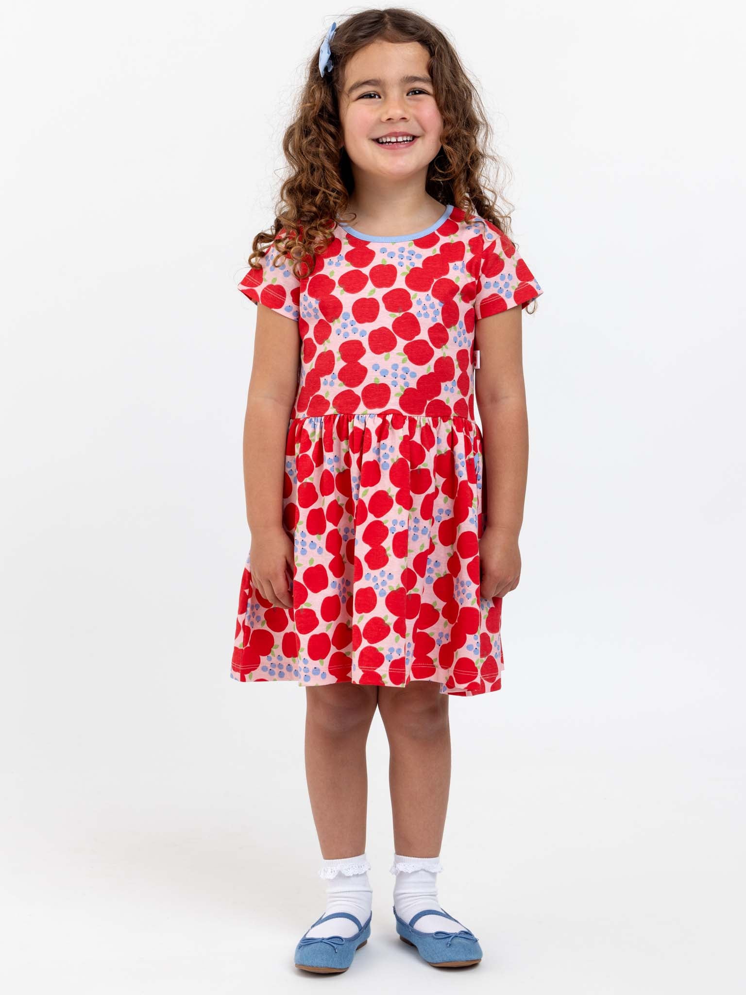 A young girl with curly hair stands smiling, wearing a Sweet Apple Amelie Dress in red and white polka dot print, white socks, and blue shoes.