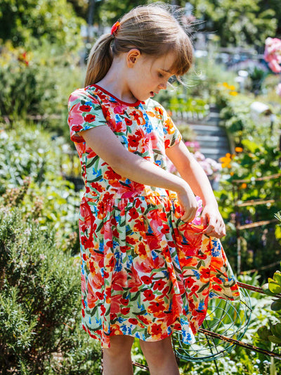 A young girl stands outdoors in a garden, admiring her attire; she is wearing the Wild Roses Amelie Dress adorned with colorful florals. Lush greenery and wild roses surround her. Her hair, tied back with a red hair tie, adds charm to her radiant presence.