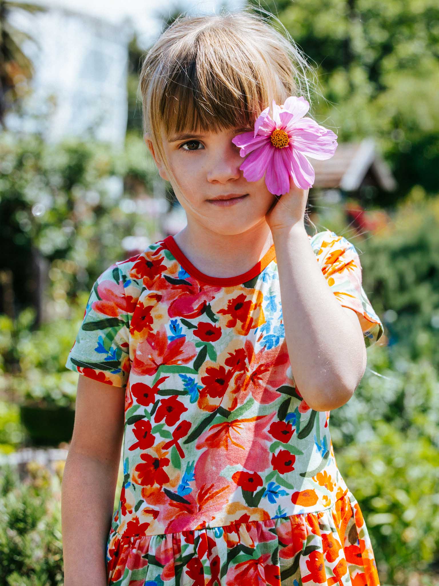 A child in the vibrant Wild Roses Amelie Dress by Wild Roses stands outdoors, holding a pink wild rose over one eye, with a blurred garden in the background.