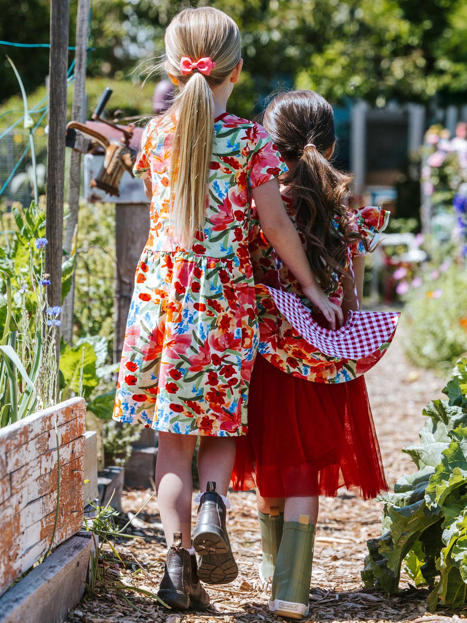 Two children in vibrant dresses and boots, one sporting the delightful Wild Roses Ariel Dress, stroll along a garden path, surrounded by lush greenery and flowers.