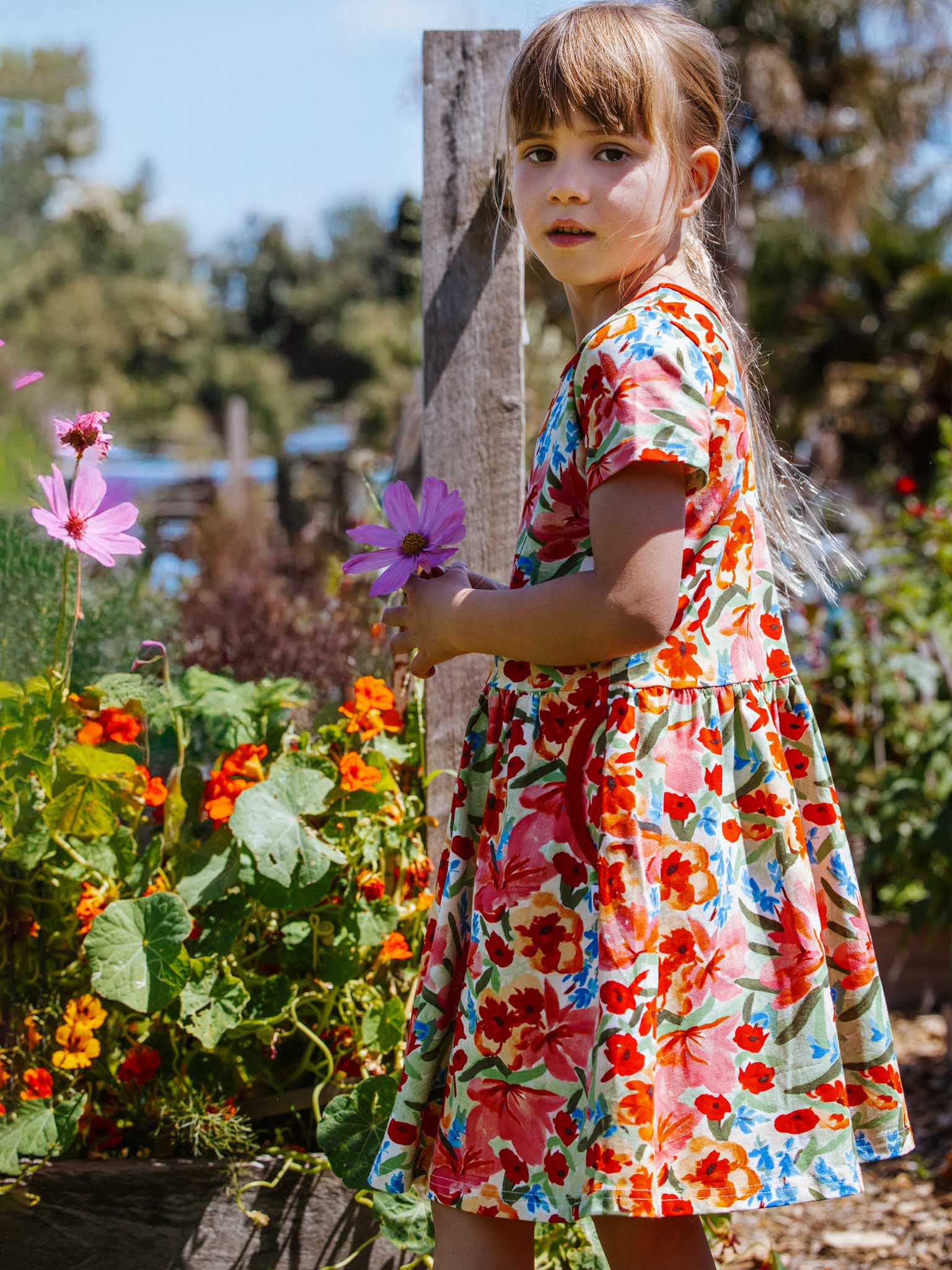 A young girl in the Wild Roses Amelie Dress, crafted from stretchy jersey, stands in a garden holding a pink flower. She is surrounded by vibrant wild roses and colorful plants amidst the trees in the background.