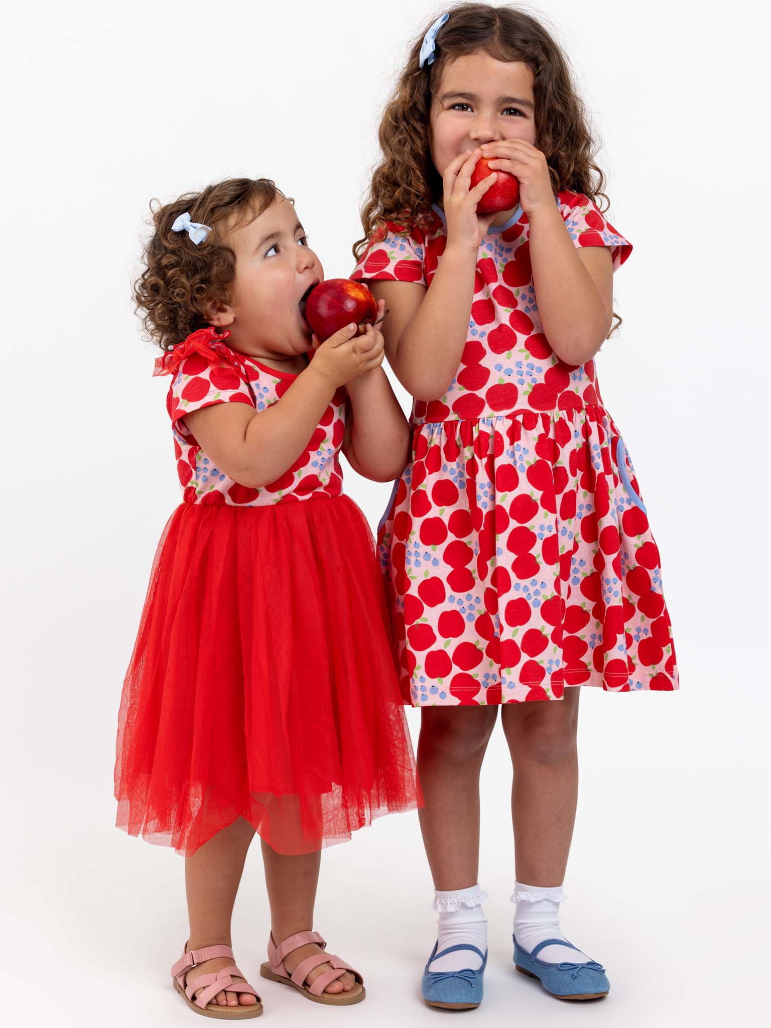 Two young girls with curly hair wearing Sweet Apple Amelie dresses stand side by side, each holding and eating a red apple. The girl on the left has a red tulle skirt; the girl on the right wears a stretchy jersey polka-dot dress.