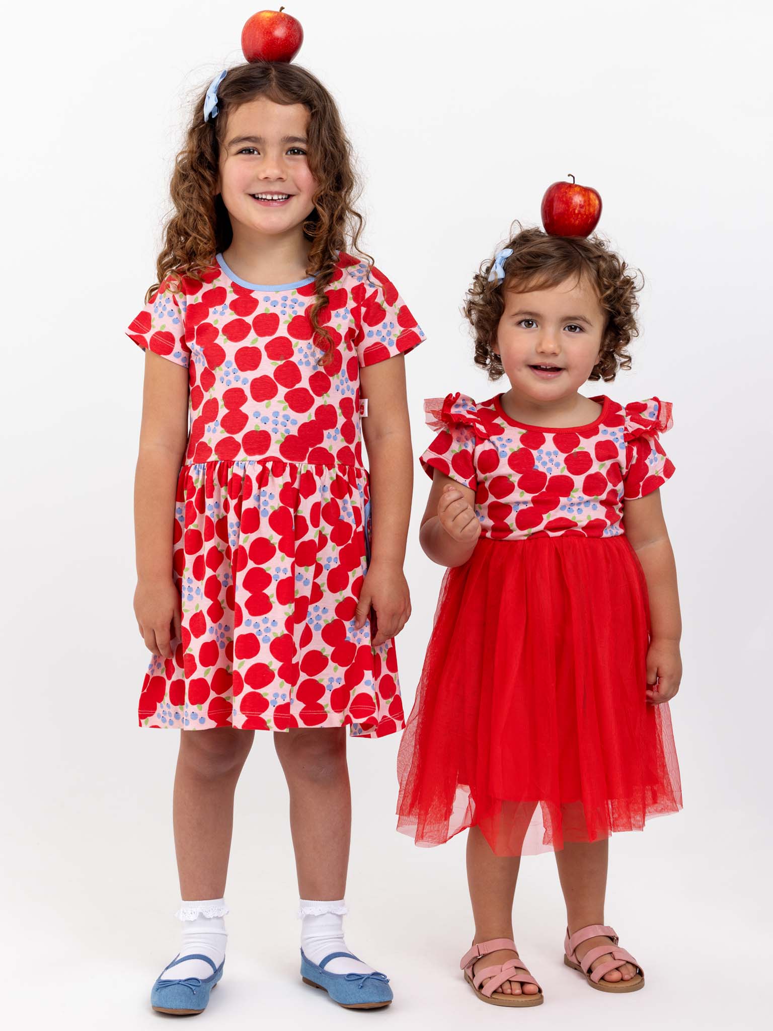 Two young girls, one taller and one shorter, are balancing red apples on their heads while wearing Sweet Apple Amelie dresses with tulle skirts. The stretchy jersey material of the Sweet Apple dresses ensures comfort, and they stand against a plain white background.