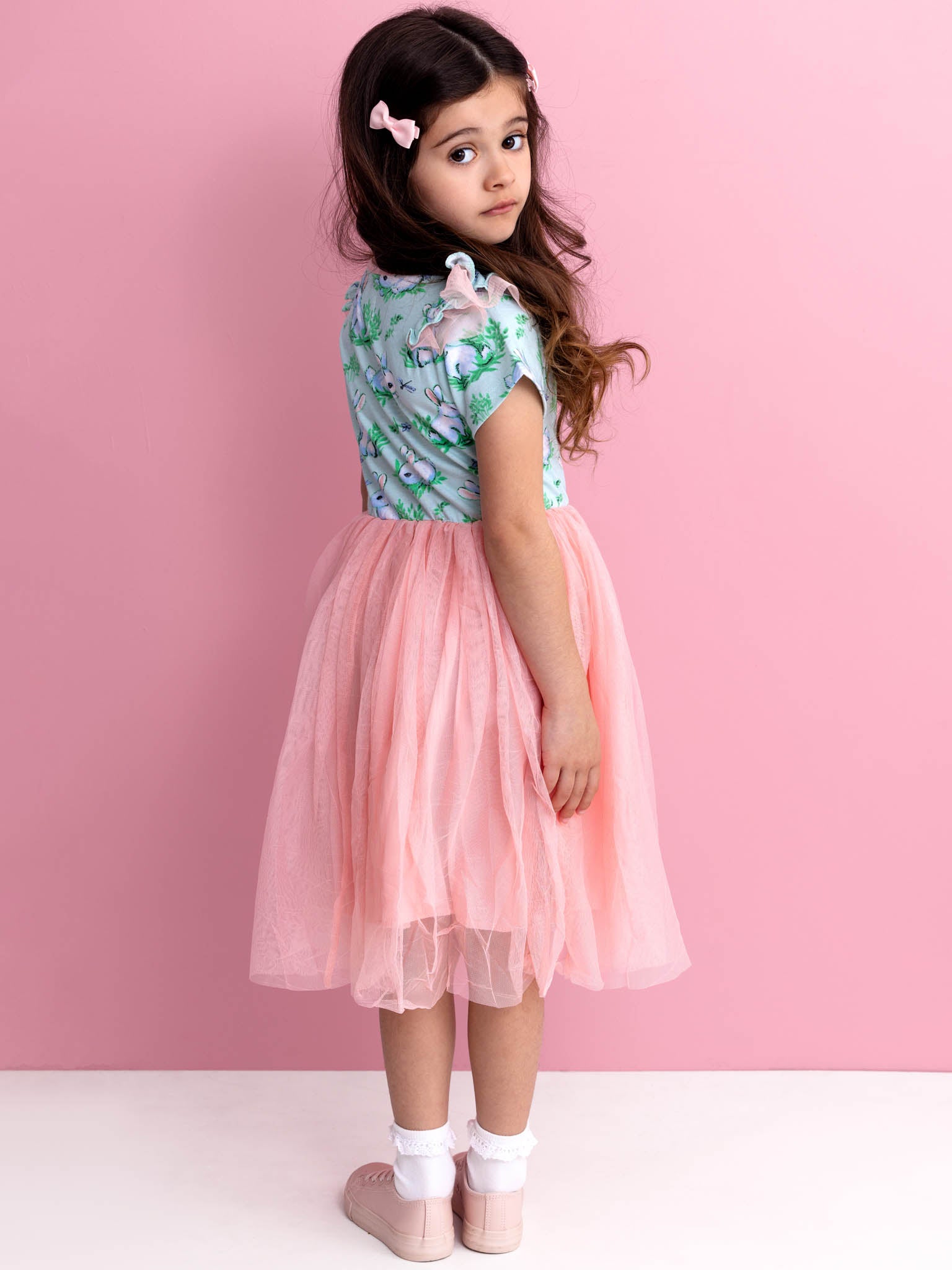 A young girl with long hair stands against a pink background, adorned in a magical Blue Bunnies Ariel Dress by Bunny. The outfit, reminiscent of adorable bunnies frolicking in a meadow, evokes the whimsy of an enchanted garden.