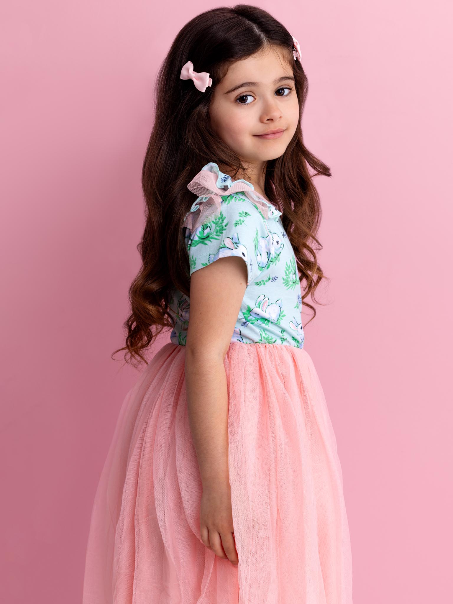 A young girl with long hair stands against a pink background, looking over her shoulder while wearing the Blue Bunnies Ariel Dress by Bunny.