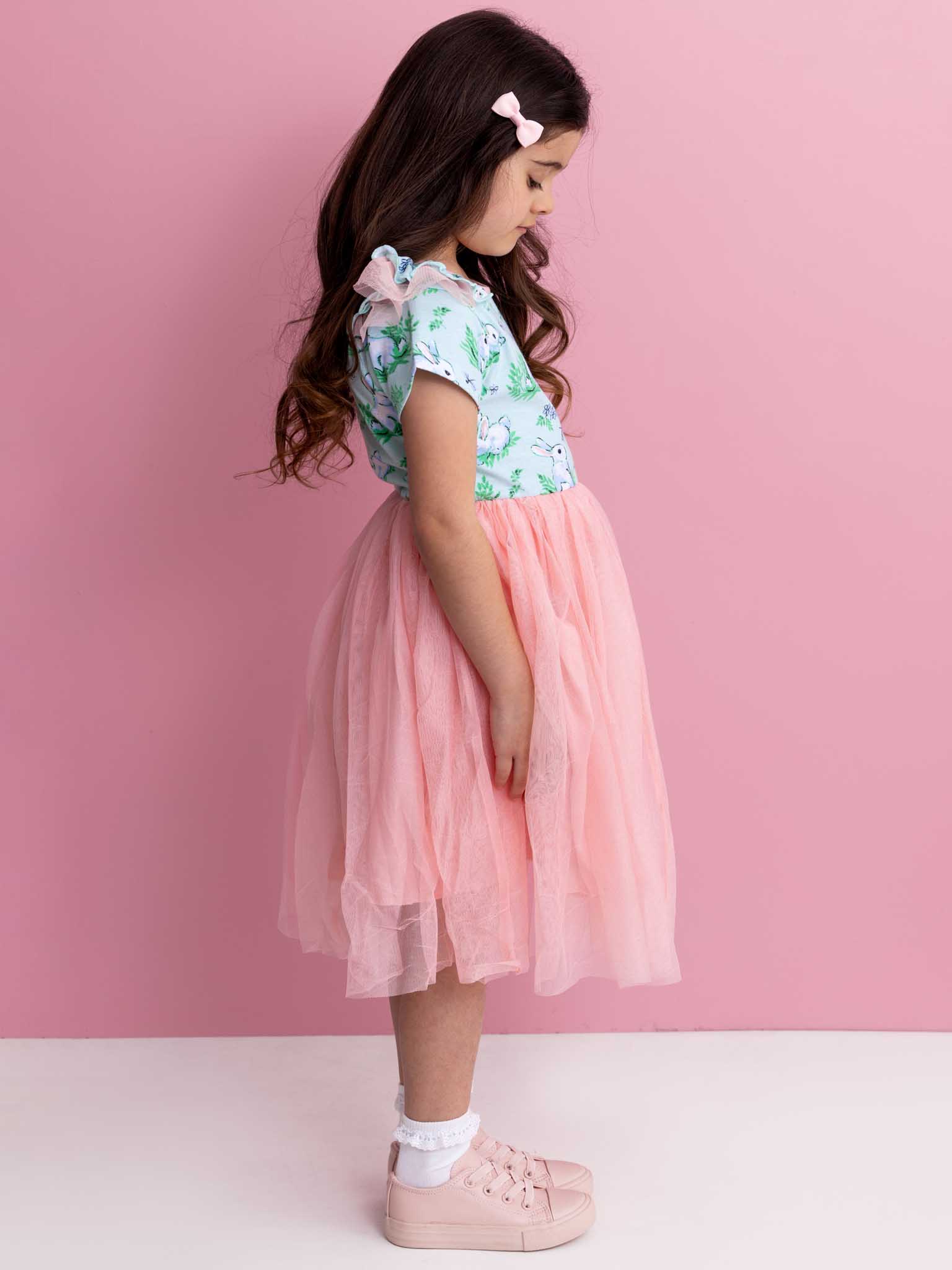 A young girl stands in profile, dressed in the Blue Bunnies Ariel Dress by Bunny. Her long hair cascades down her back, complemented by pink shoes, all set against a soft pink background.