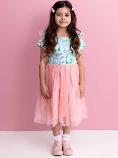 A young girl stands in front of a pink background, dressed in the charming Blue Bunnies Ariel Dress by Bunny. Her look is completed with pink hair clips and adorable bunny-shaped shoes.