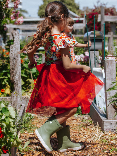 A young girl in a red Wild Roses Ariel Dress and green boots walks in a garden adorned with wild roses, as another child plays nearby. Flowers and plants surround them, creating a serene and enchanting scene.