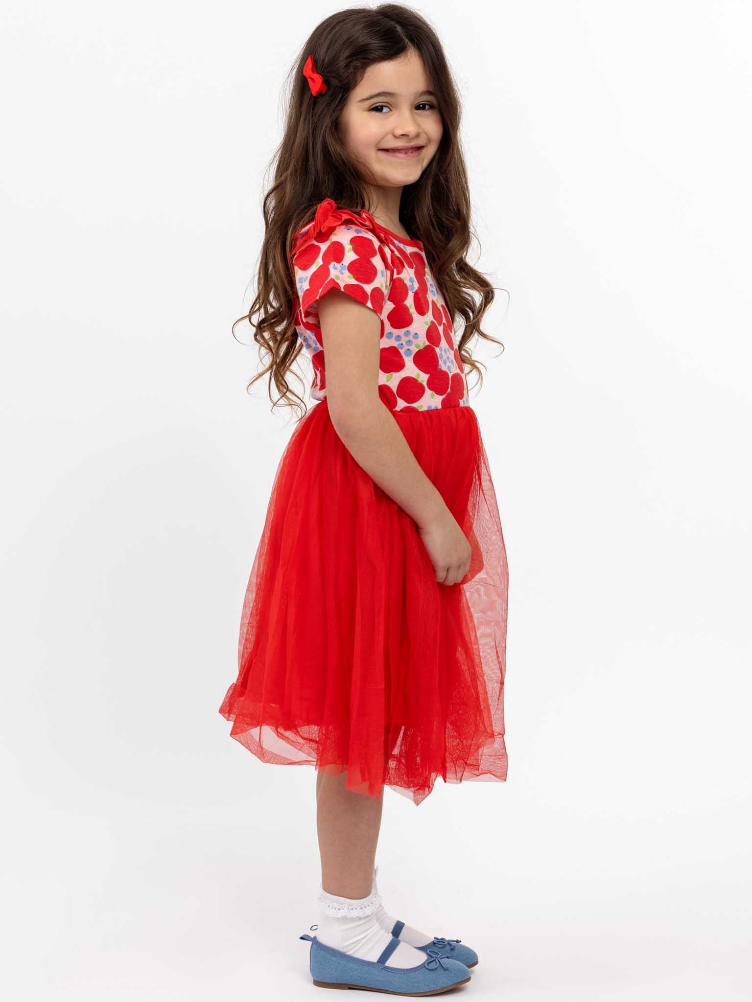 A young girl with long brown hair is wearing the Sweet Apple Ariel Dress by Sweet Apple, featuring a stretch jersey bodice and a red tulle skirt. She stands against a white background, smiling with a red bow in her hair and blue shoes, radiating pure tutu goodness.