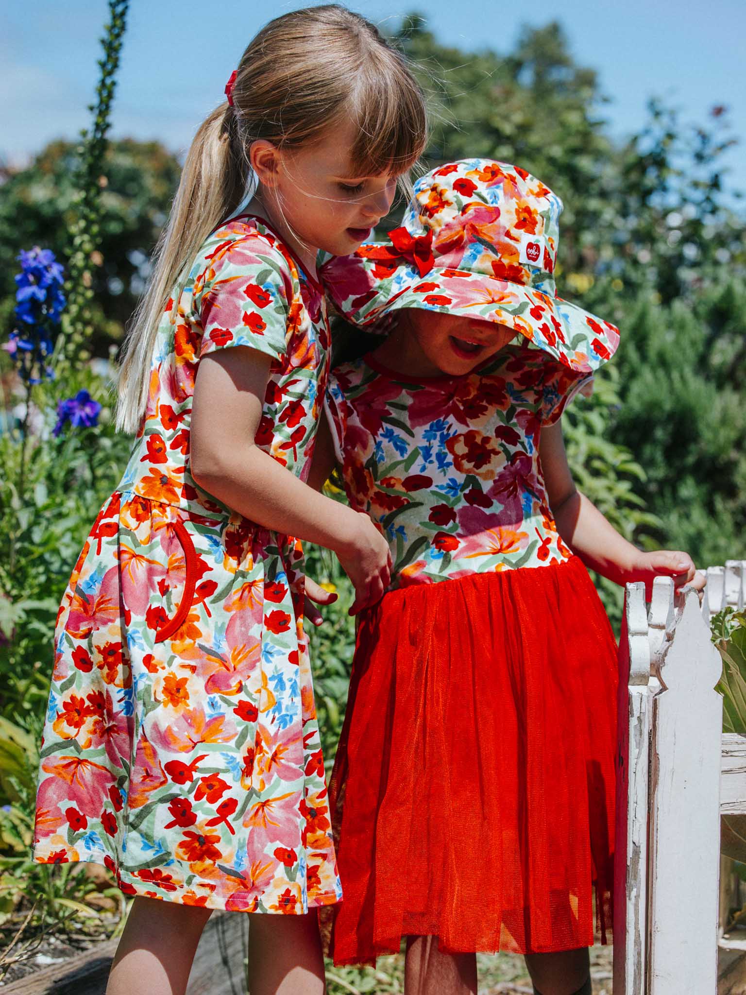 Two young girls in vibrant Wild Roses Ariel Dresses and sun hats stand gracefully in a garden next to a wooden fence.