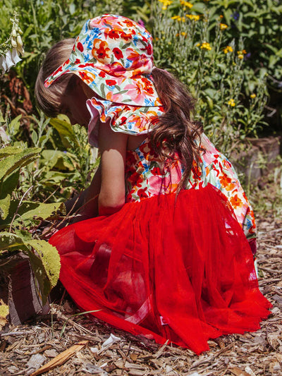 In a garden setting, a child wearing a floral hat and the Wild Roses Ariel Dress with a red skirt kneels, tending to plants. The delicate print of the Wild Roses brand dress complements the scene beautifully.