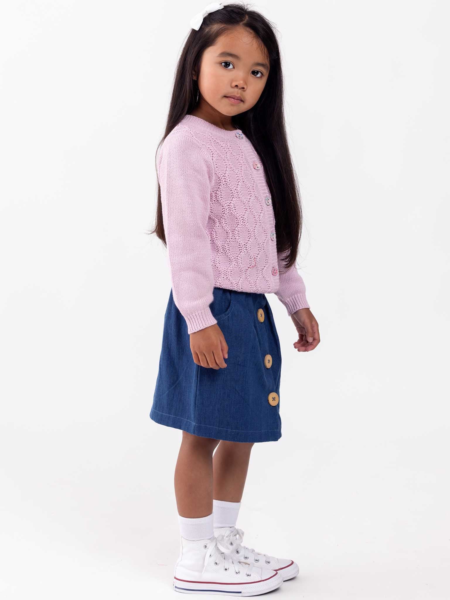 A young girl in a Audrina Violet Cardigan from Knitwear stands with one hand on her hip, looking to the side against a white background.