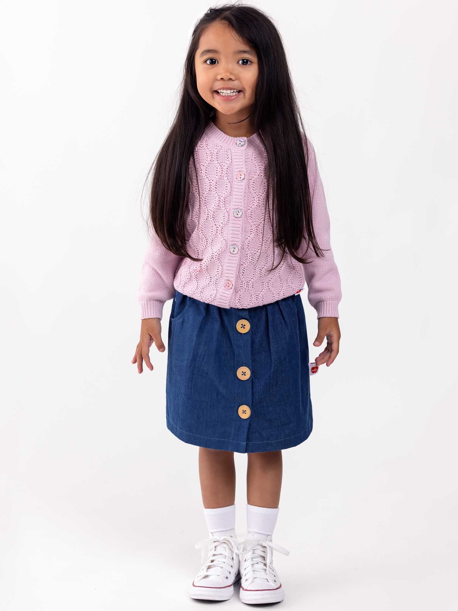 A young girl in a pink Audrina Violet Cardigan by Knitwear, blue skirt, and white sneakers stands smiling against a plain background.