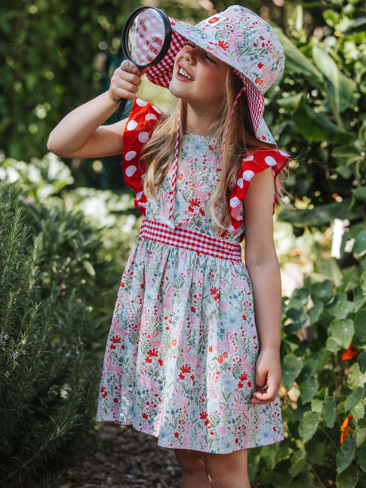 A young girl in a floral dress and a wide brim Bunny hat with a ponytail hole is holding a magnifying glass up to her eye in a garden.