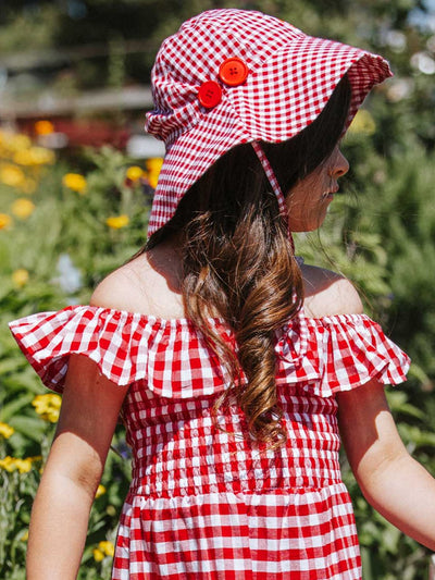 In a blooming garden full of yellow flowers, a child gazes to the side wearing a Red Gingham checkered dress made from 100% cotton, complemented by a matching Red Gingham Bucket Hat accented with red buttons.