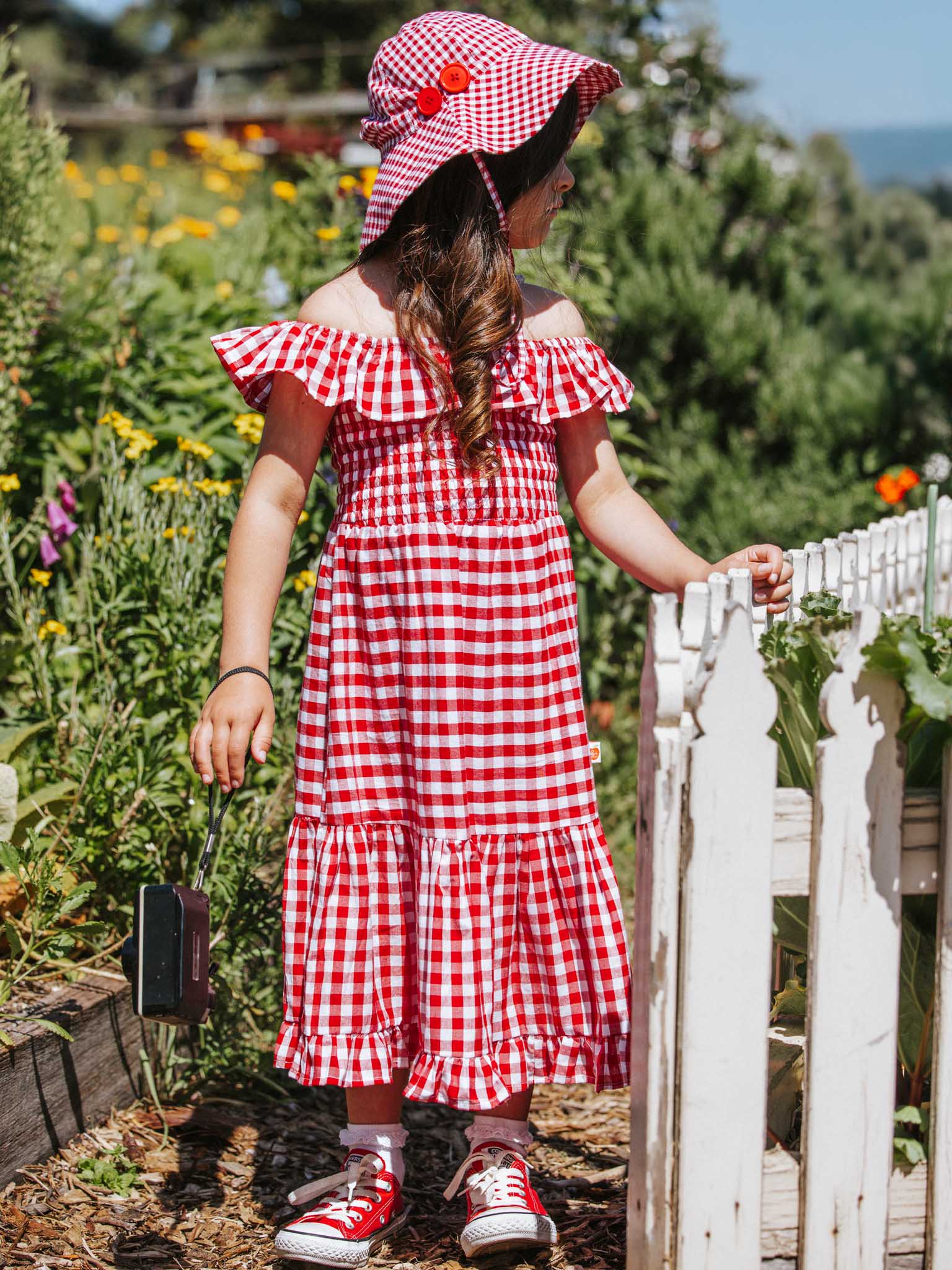 A girl wearing a Red Gingham bucket hat and matching hand-printed style red and white checkered dress stands by a white fence, holding a small black object. She is surrounded by lush greenery and colorful flowers, creating a picturesque scene.